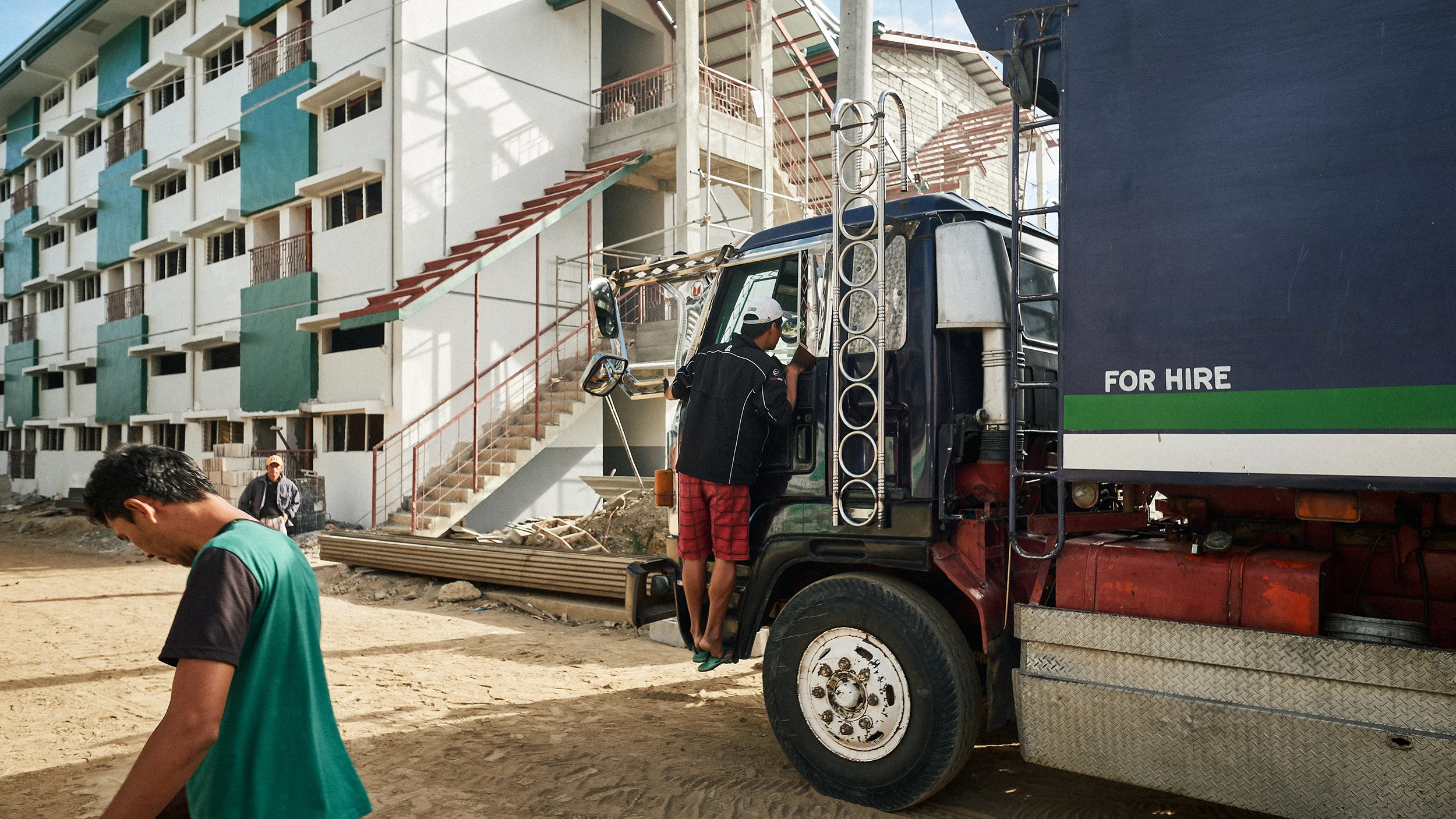 Construction site with a truck labeled 'for hire,' a person climbing into the truck, and a partially constructed building in the background.