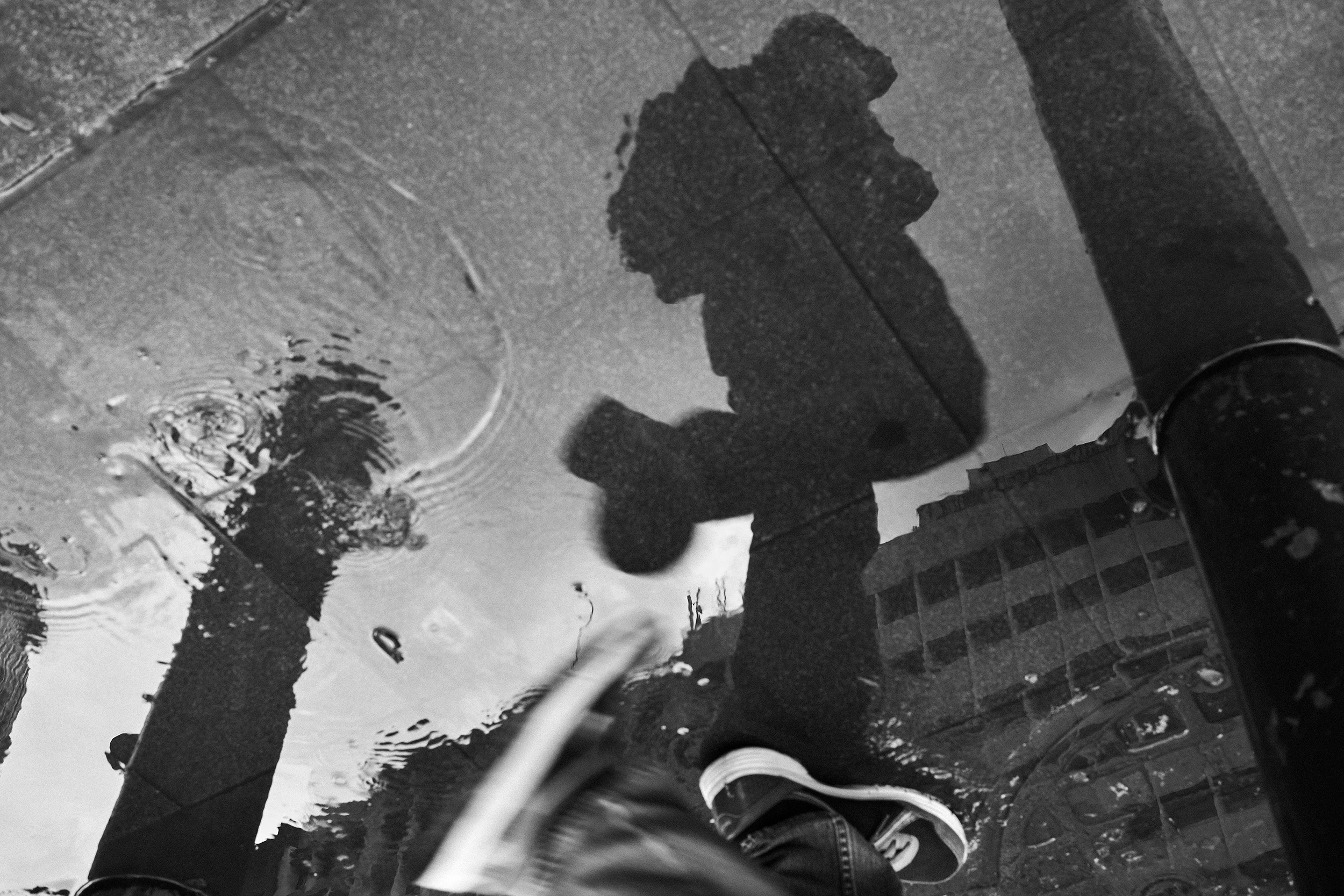 Reflection of a person walking in a puddle on a city street with visible buildings and poles.