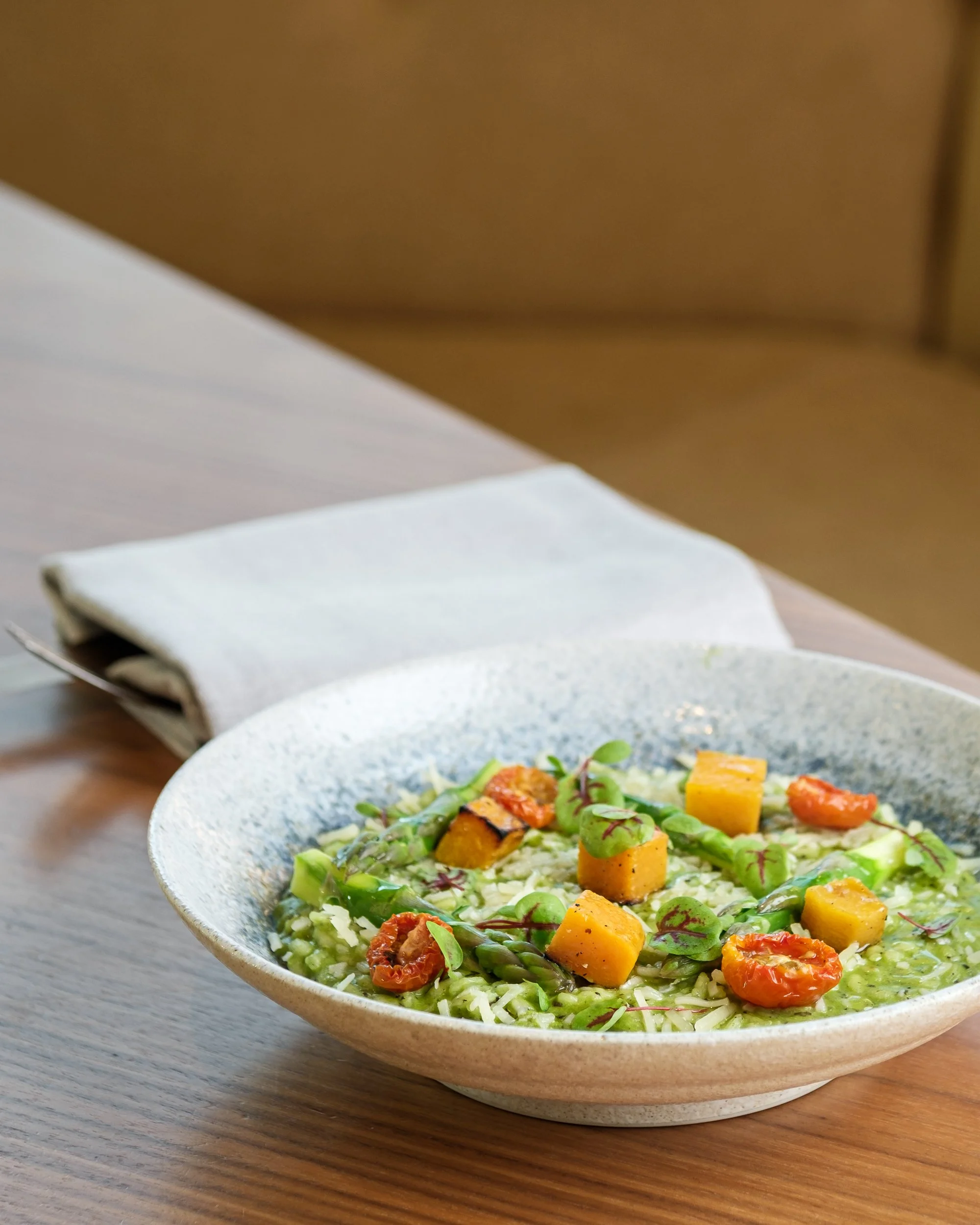 A bowl of pasta topped with cherry tomatoes, yellow cheese cubes, asparagus, and greens on a wooden table, with a folded napkin nearby.