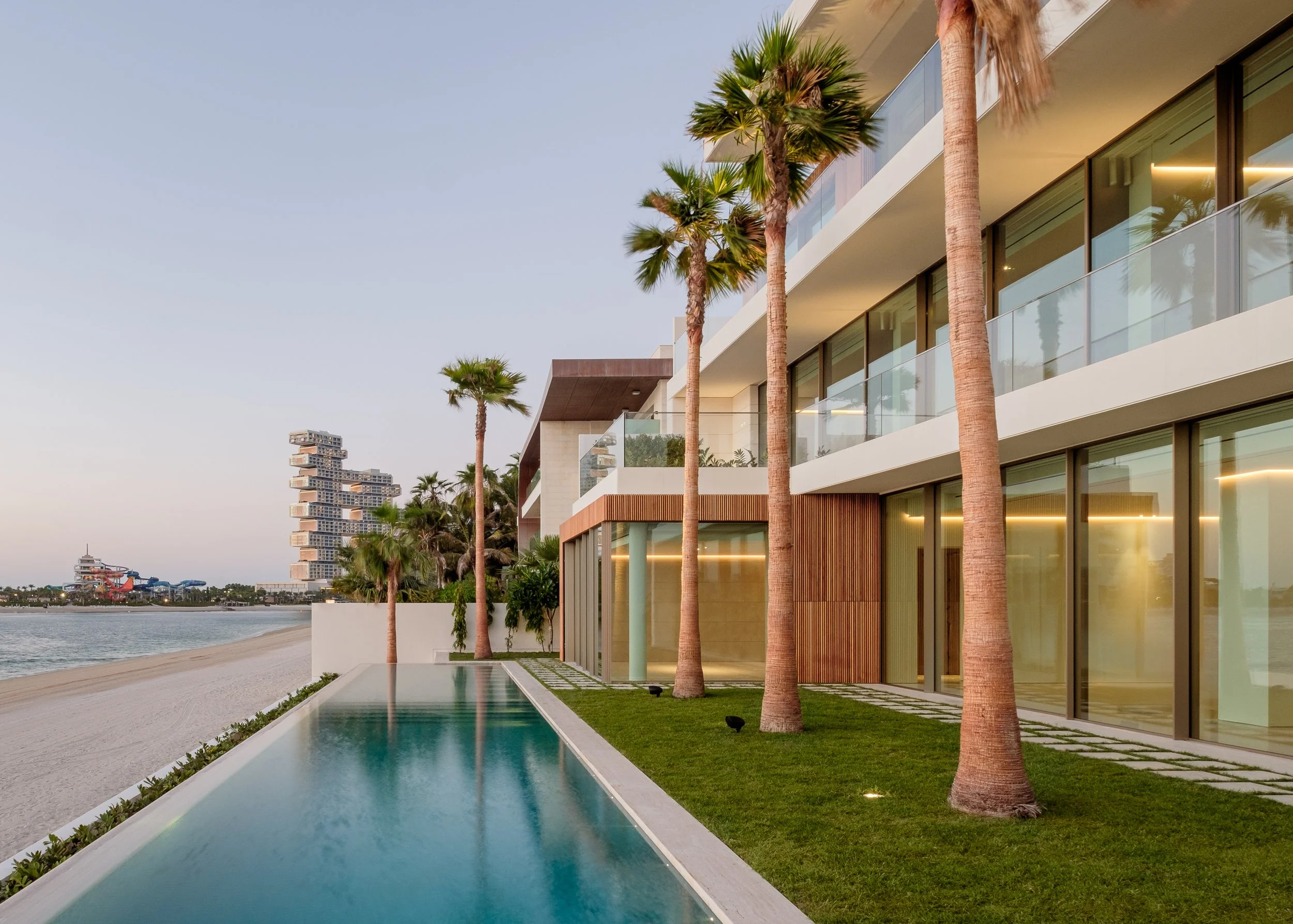 Modern beachside hotel with pool, palm trees, and a cityscape in the background.