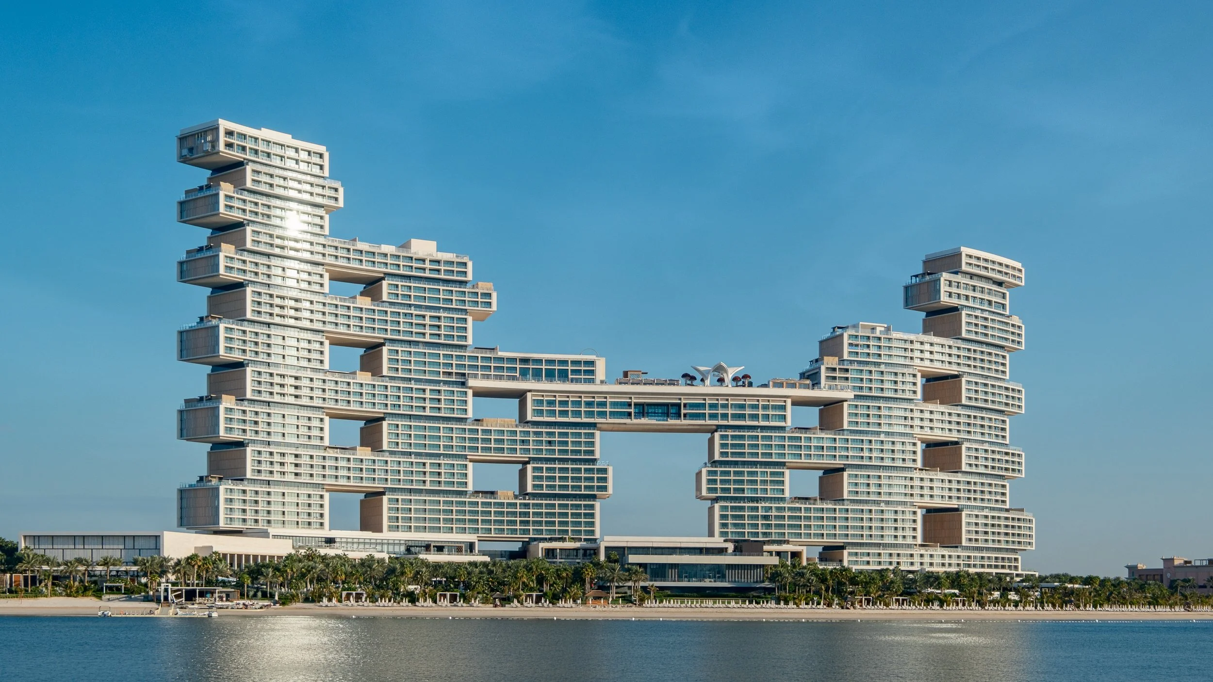 Modern high-rise building with staggered, stacked design and glass balconies, set against a clear blue sky, with water and palm trees in the foreground.