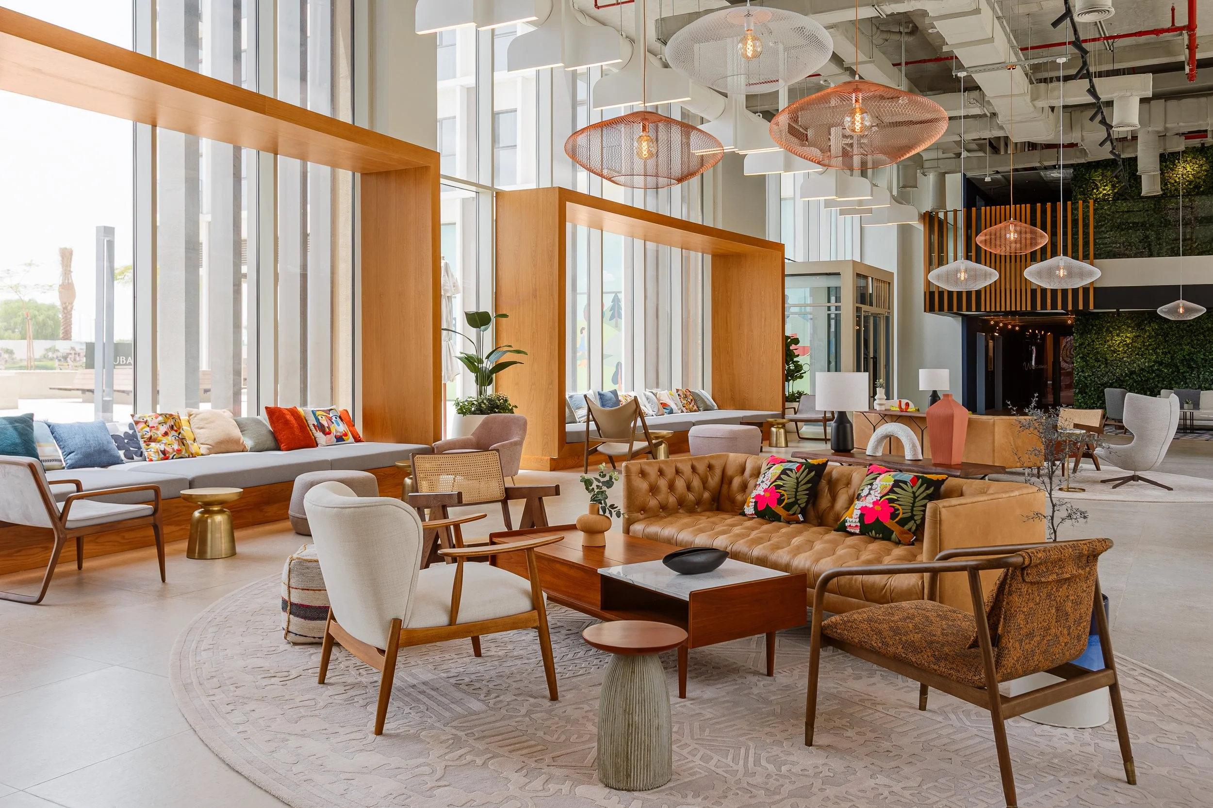 Modern, stylish indoor lounge with large windows, wooden accents, colorful pillows, and a variety of chairs and sofas, including a leather tufted sofa, with decorative lamps and hanging light fixtures.