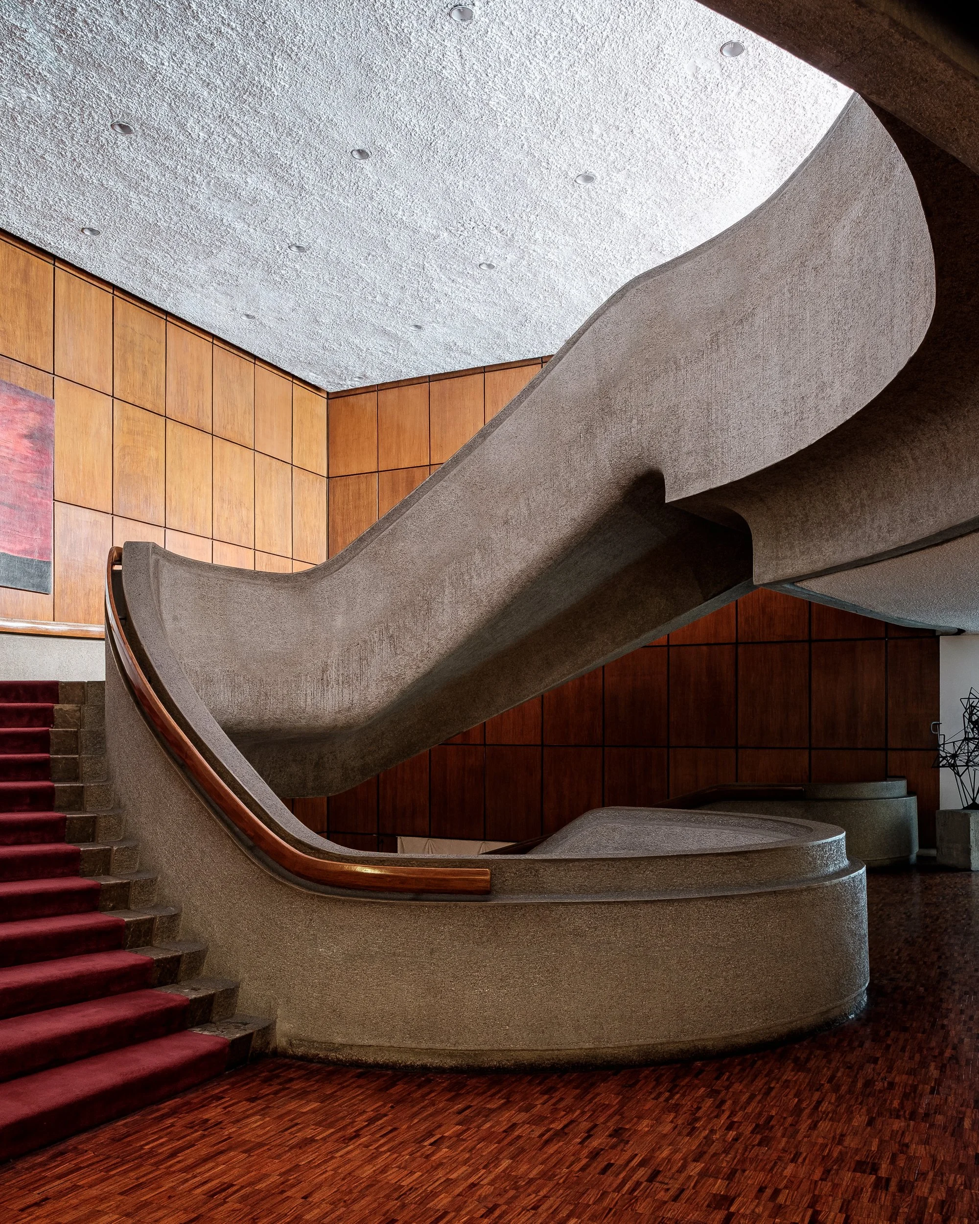 Interior view of a building featuring high curved concrete staircase with wooden railings, walls covered with wood panels, a patterned red carpet on stairs, and a textured ceiling with small recessed lights.