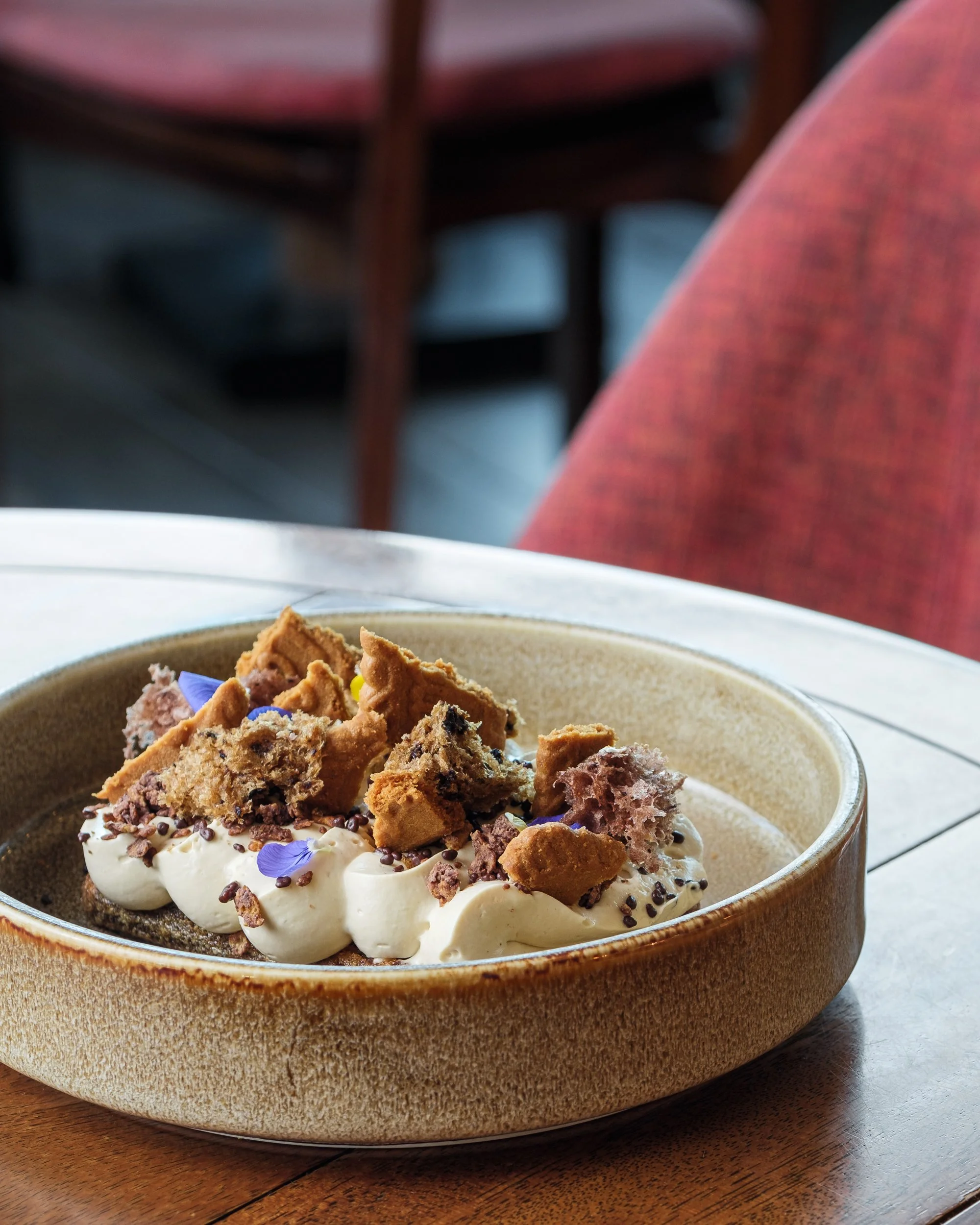 Dessert dish with whipped cream, chocolate crumbs, and broken biscuit pieces, garnished with purple edible flowers in a brown bowl on a wooden table.