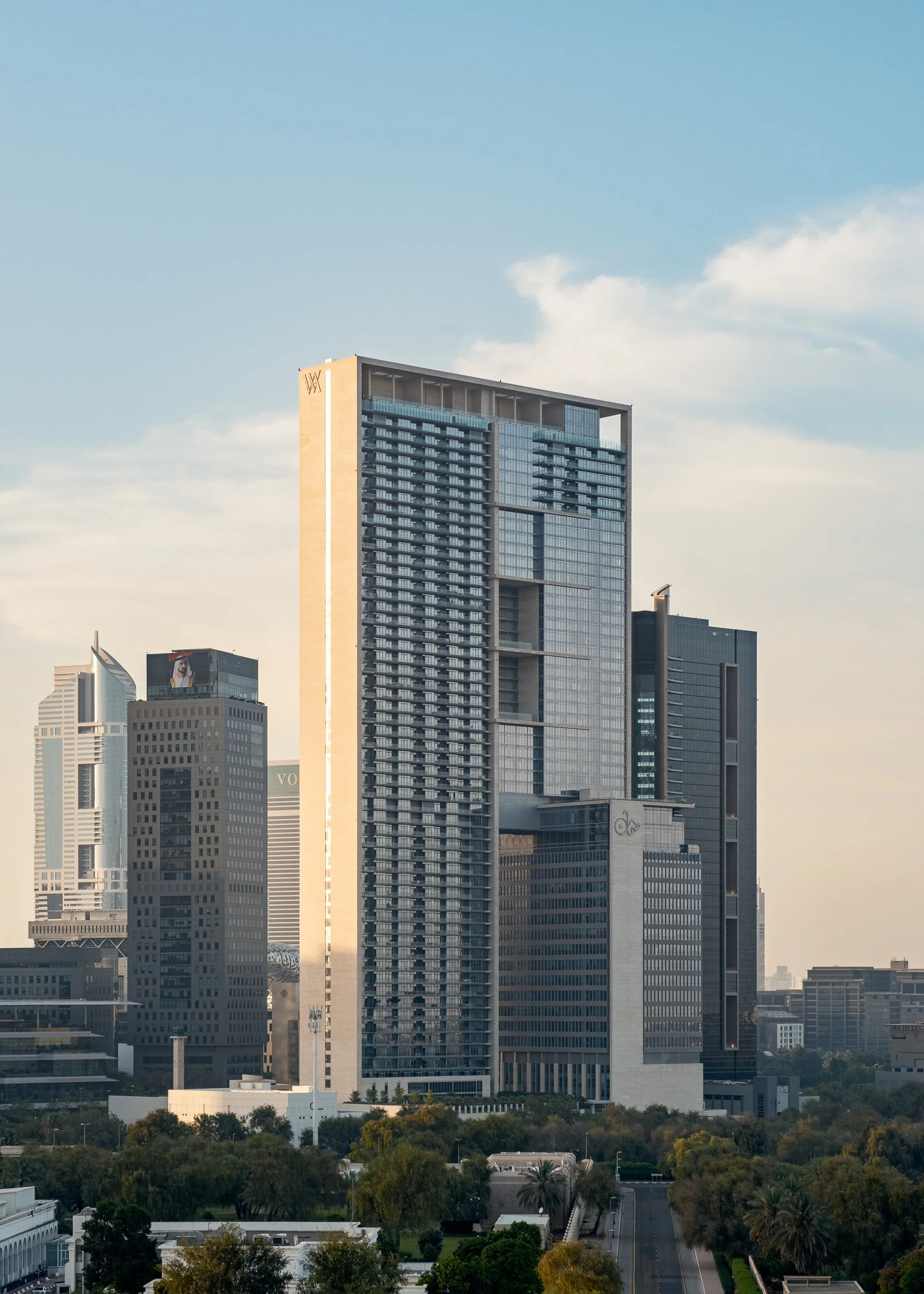 Skyline with high-rise skyscraper and neighboring buildings, trees in the foreground, clear sky with a few clouds.