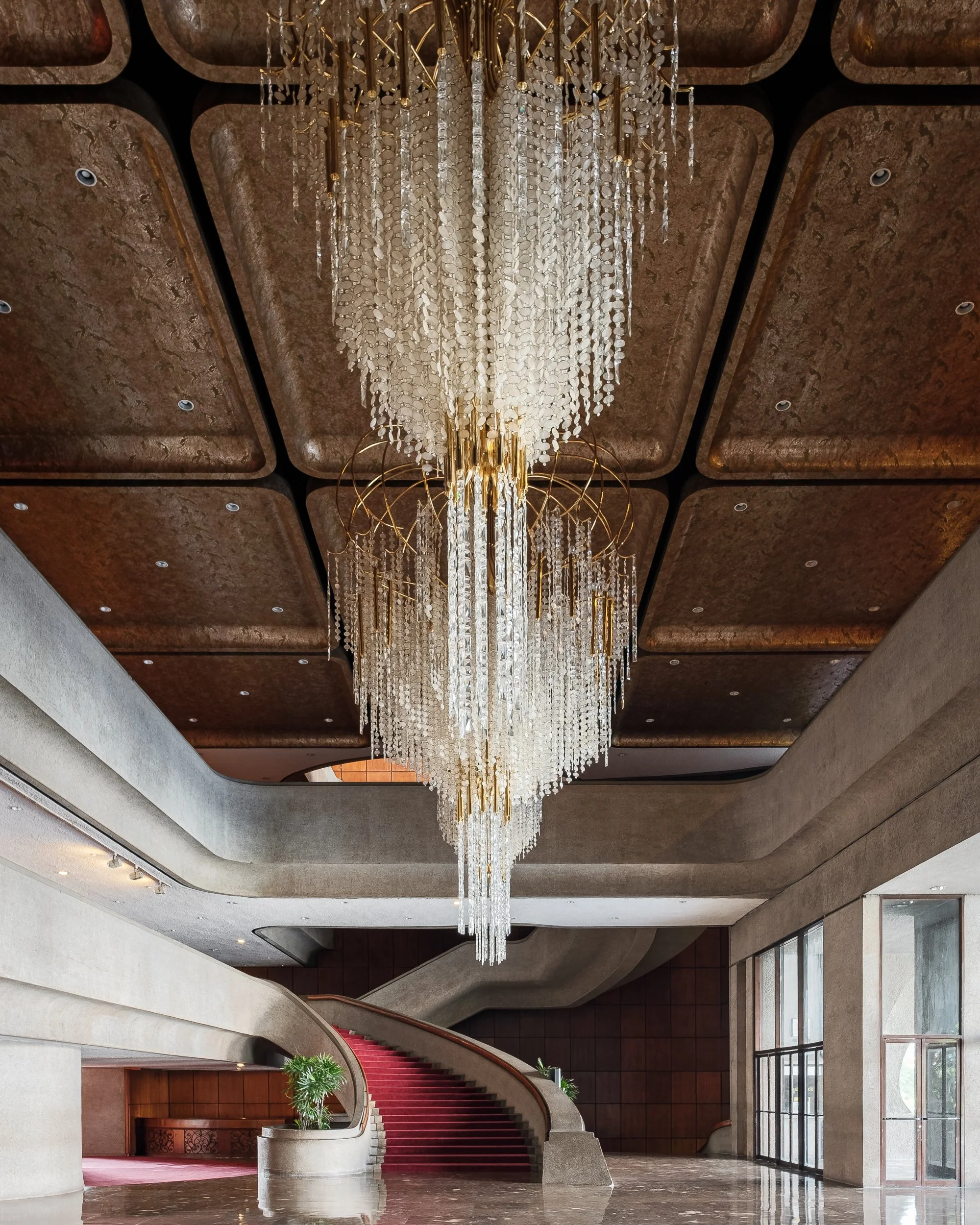 Elegant hotel lobby with a large crystal chandelier hanging from the ceiling, a curved staircase with a red carpet runner, and a marble floor.