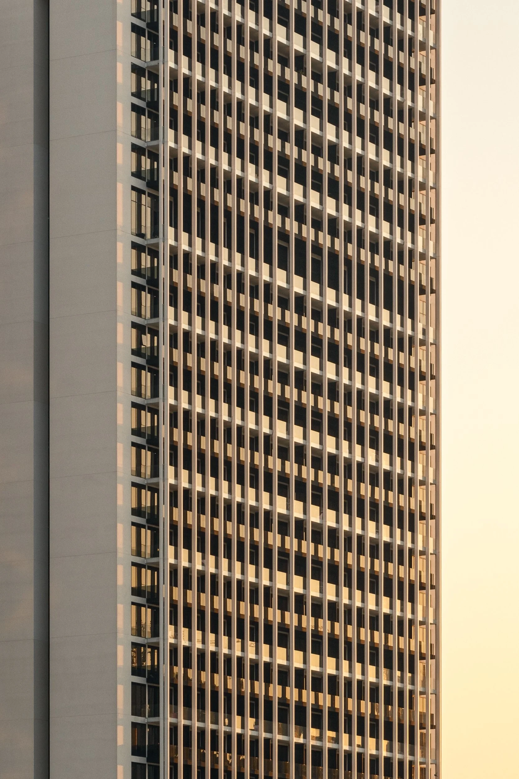 Close-up of a modern high-rise building with a repetitive geometric pattern of windows and balconies, bathed in warm sunset light.
