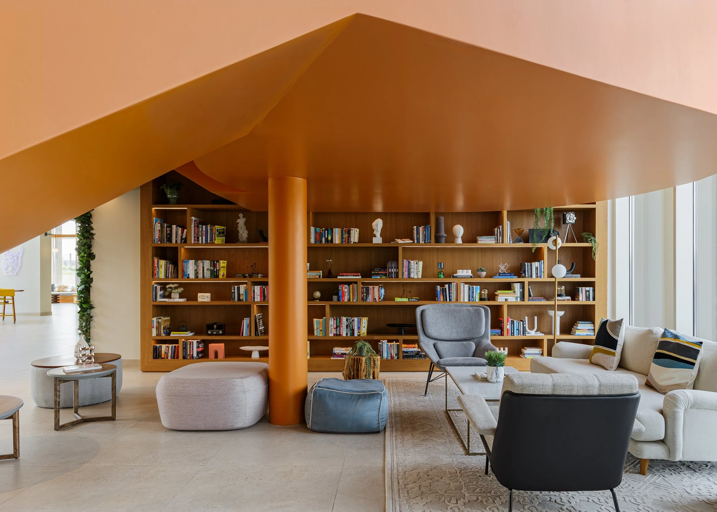 Modern hotel or library lounge area with a wooden bookshelf, comfortable chairs, a beige sofa, and a large orange ceiling structure.