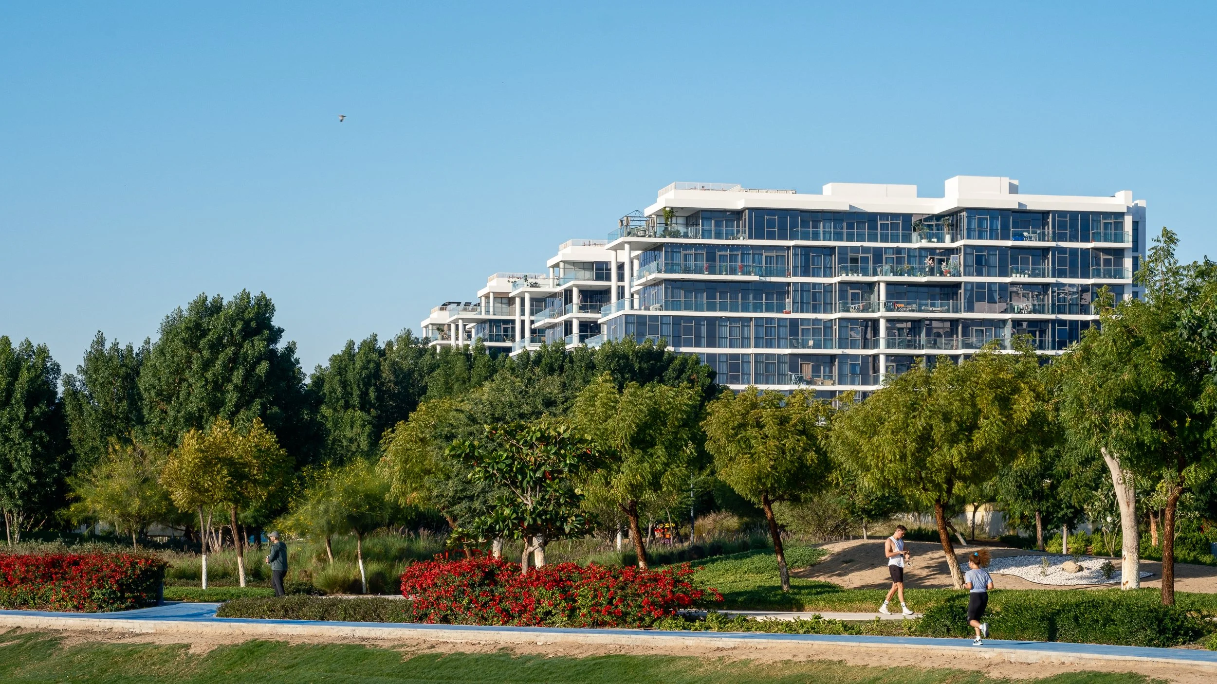 Modern multi-story apartment building with glass balconies, surrounded by lush green trees and a park with people walking.