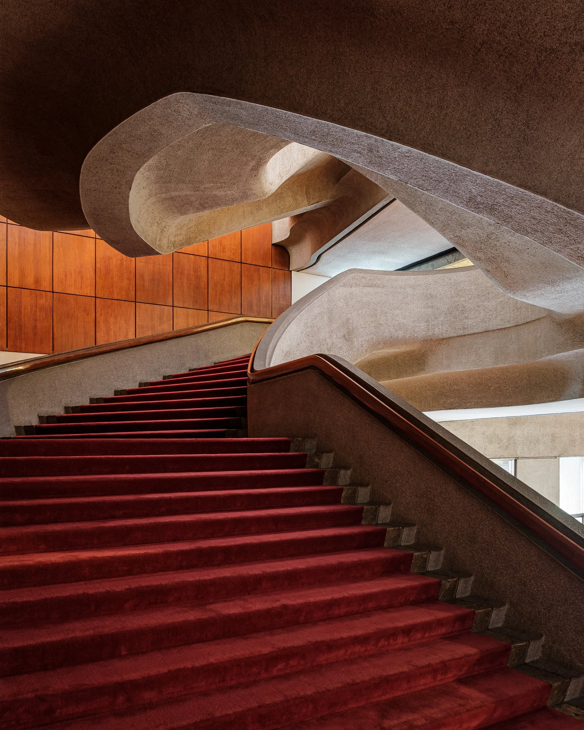 Indoor staircase with red carpet, curved beige walls, and wooden paneling on the floor.