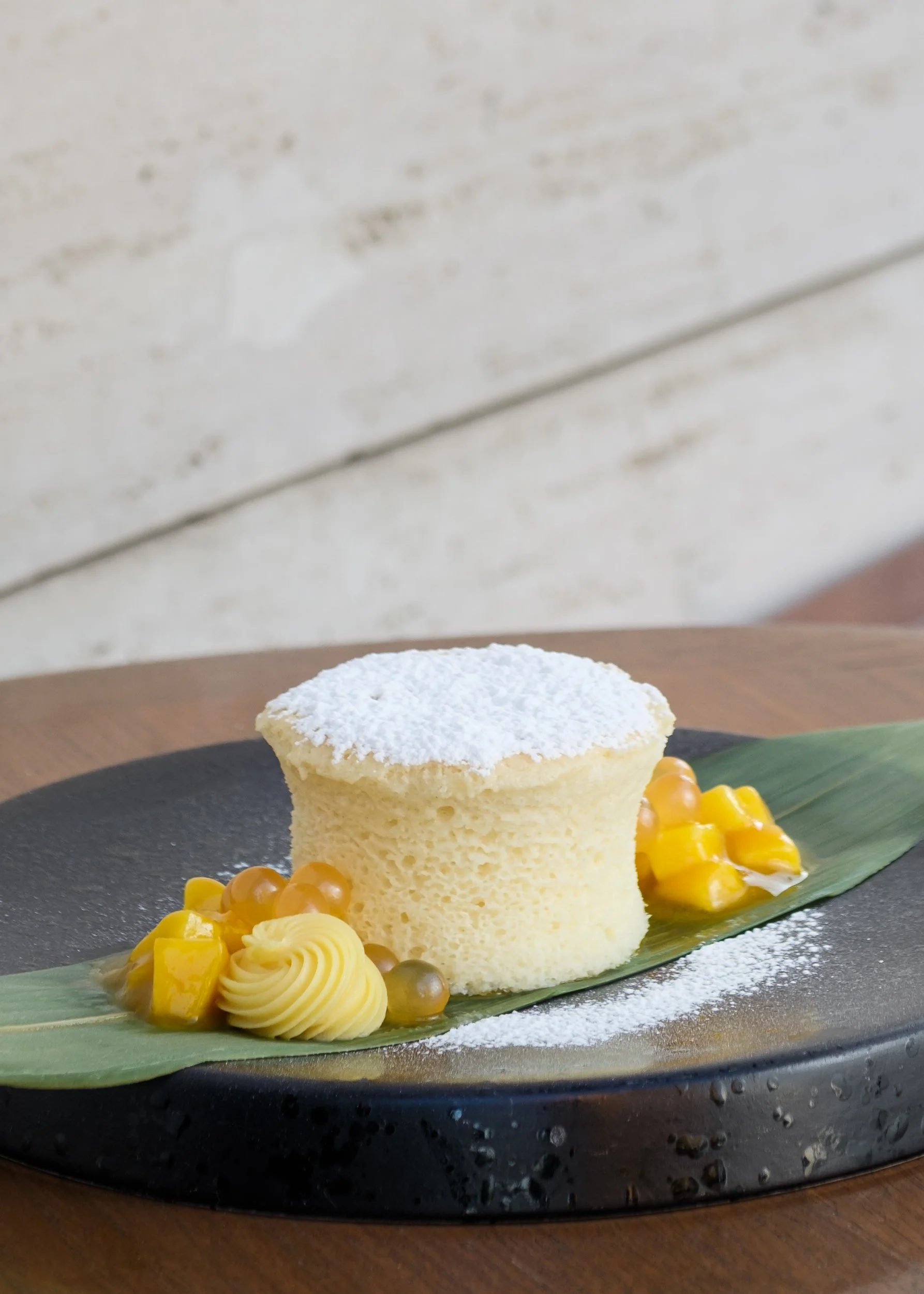 A piece of sponge cake dusted with powdered sugar, served with whipped cream, mango chunks, and orange tapioca pearls on a leaf, all placed on a black plate.