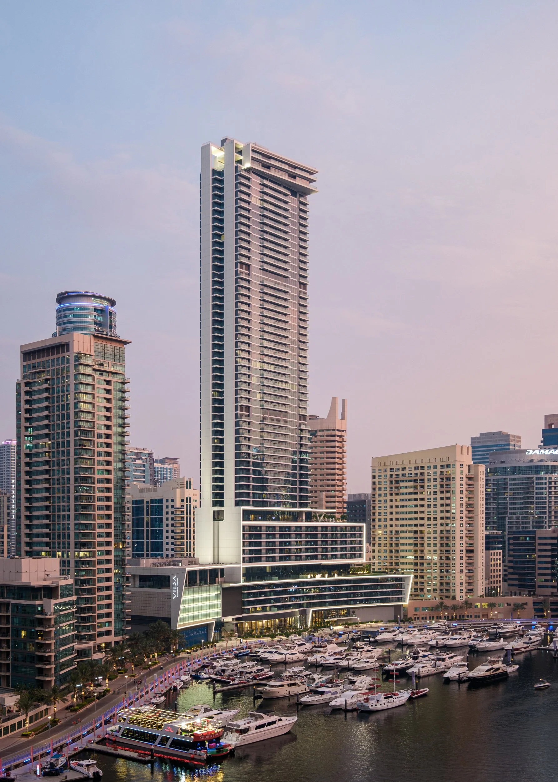 Skyline view of tall modern skyscrapers near a marina with yachts, with city lights beginning to turn on as evening approaches.