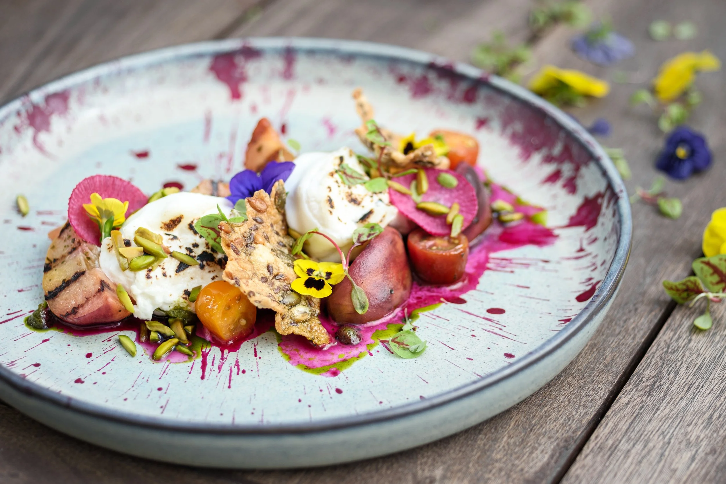 A gourmet dessert on a ceramic plate with grilled watermelon, cherry tomatoes, mint leaves, edible flowers, seeds, and a dollop of cream, garnished with colorful flower petals and edible flowers.