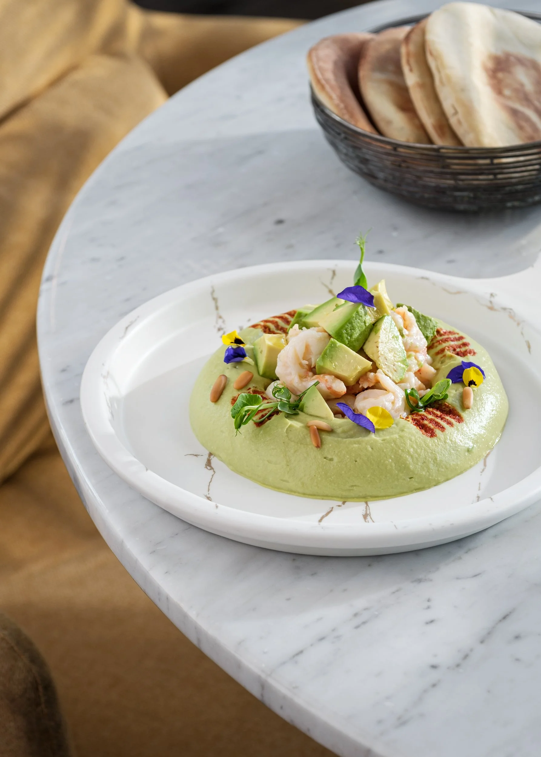 A gourmet dish with green sauce topped with shrimp, avocado, and edible flowers, served on a white marble table.
