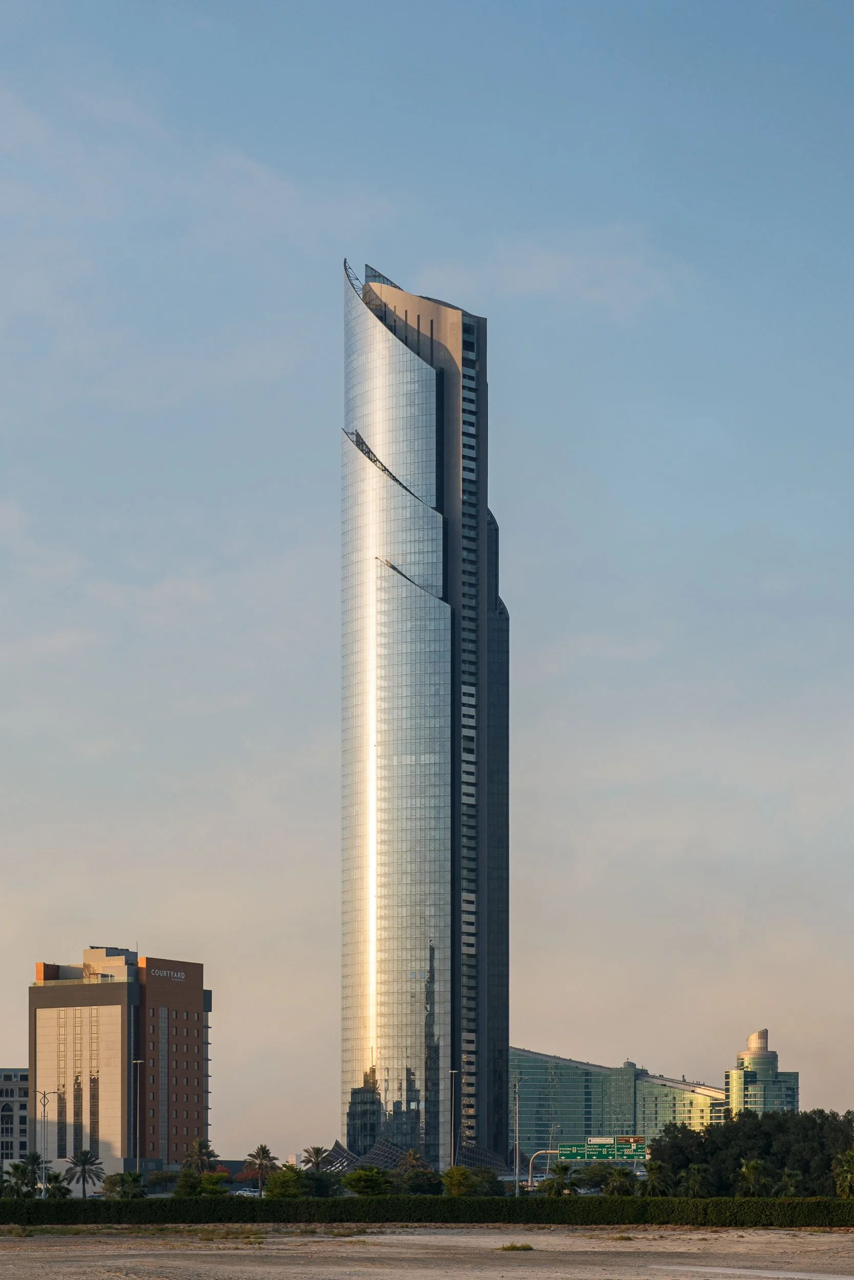 A modern skyscraper with a curved glass facade and a distinctive peaked top, set against a clear blue sky with a few clouds. Surrounding buildings are also visible in the foreground.