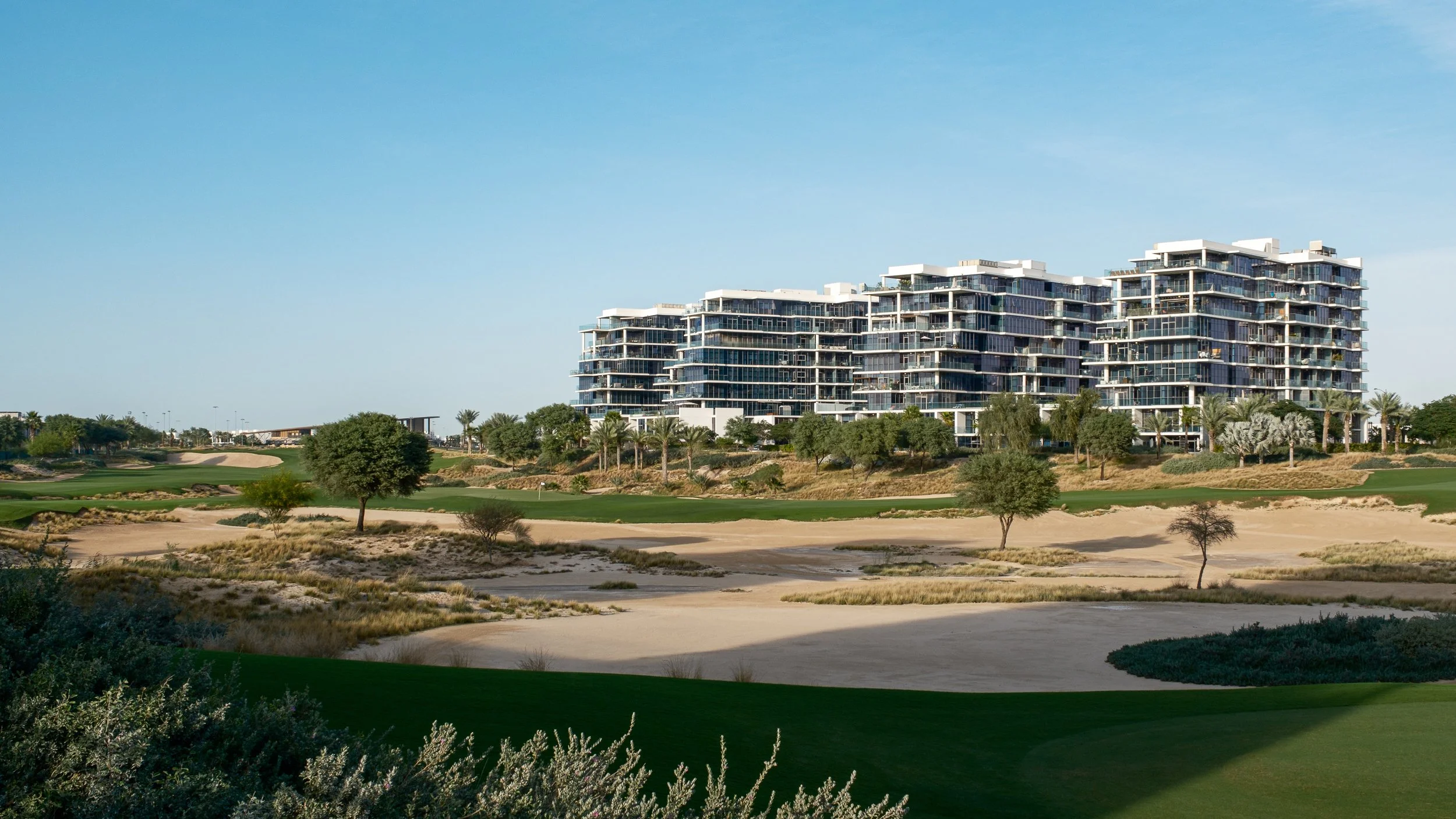Modern multi-story residential building with glass balconies, surrounded by a landscaped area with trees, bushes, and a sandy golf course, under a clear blue sky.