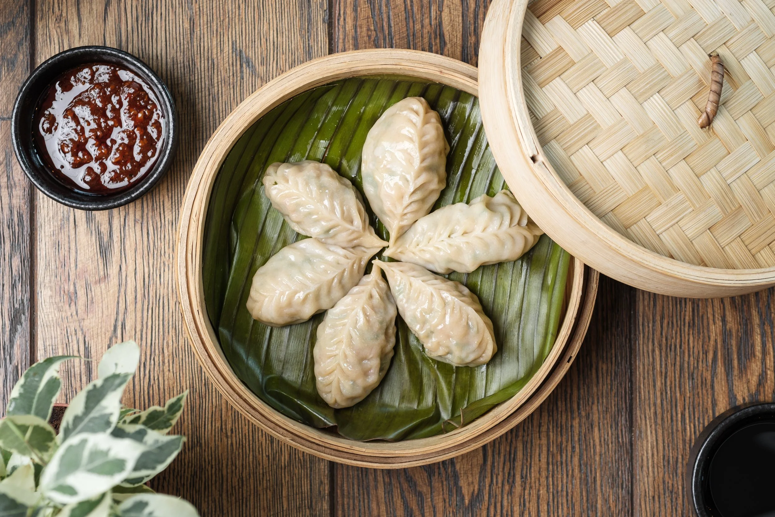 Dumplings arranged in a circle on a banana leaf inside a bamboo steamer, with dipping sauces on the side.