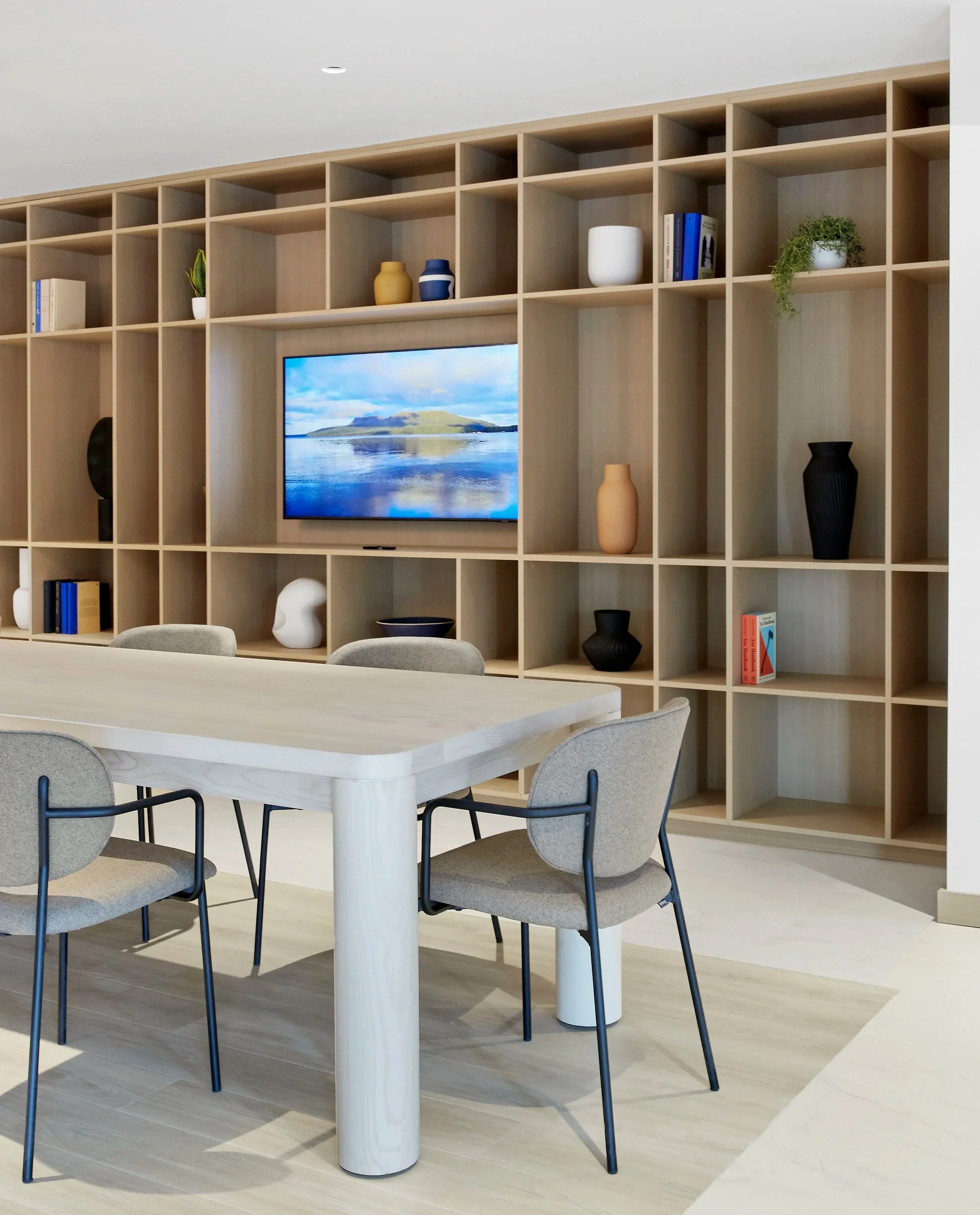 Co-workspace at Station No. 3 Condo in Whitby, shelving unit with decorative objects and books, a wall-mounted flat-screen TV, a light-coloured conference table, with upholstered chairs.