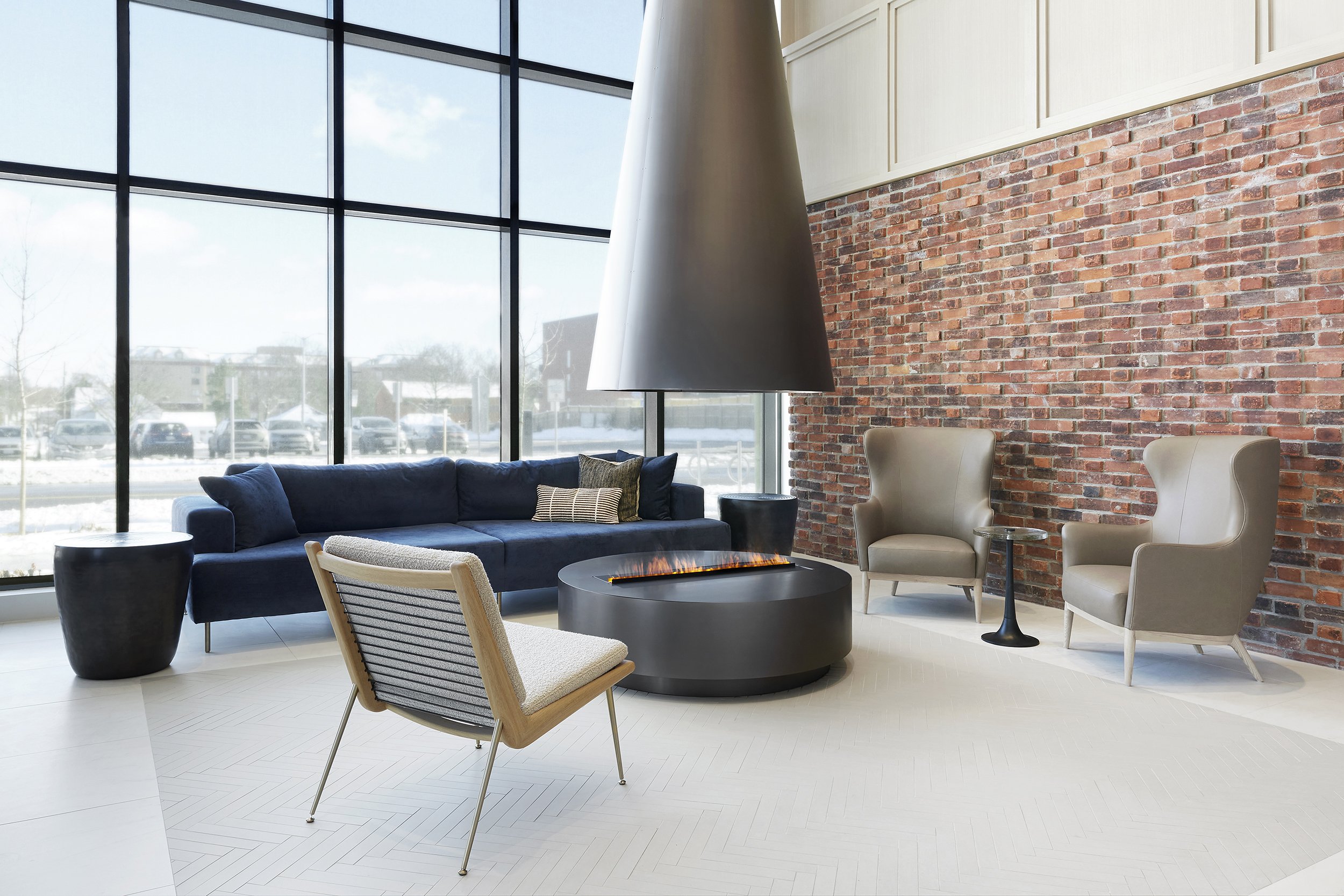 Modern lobby lounge at Station No. 3 Condominiums in Whitby by Brookfield Properties, a restored historic fire station with large windows, street views, a blue sofa, armchairs, a circular  fireplace with an overhead hood, and an exposed brick wall.