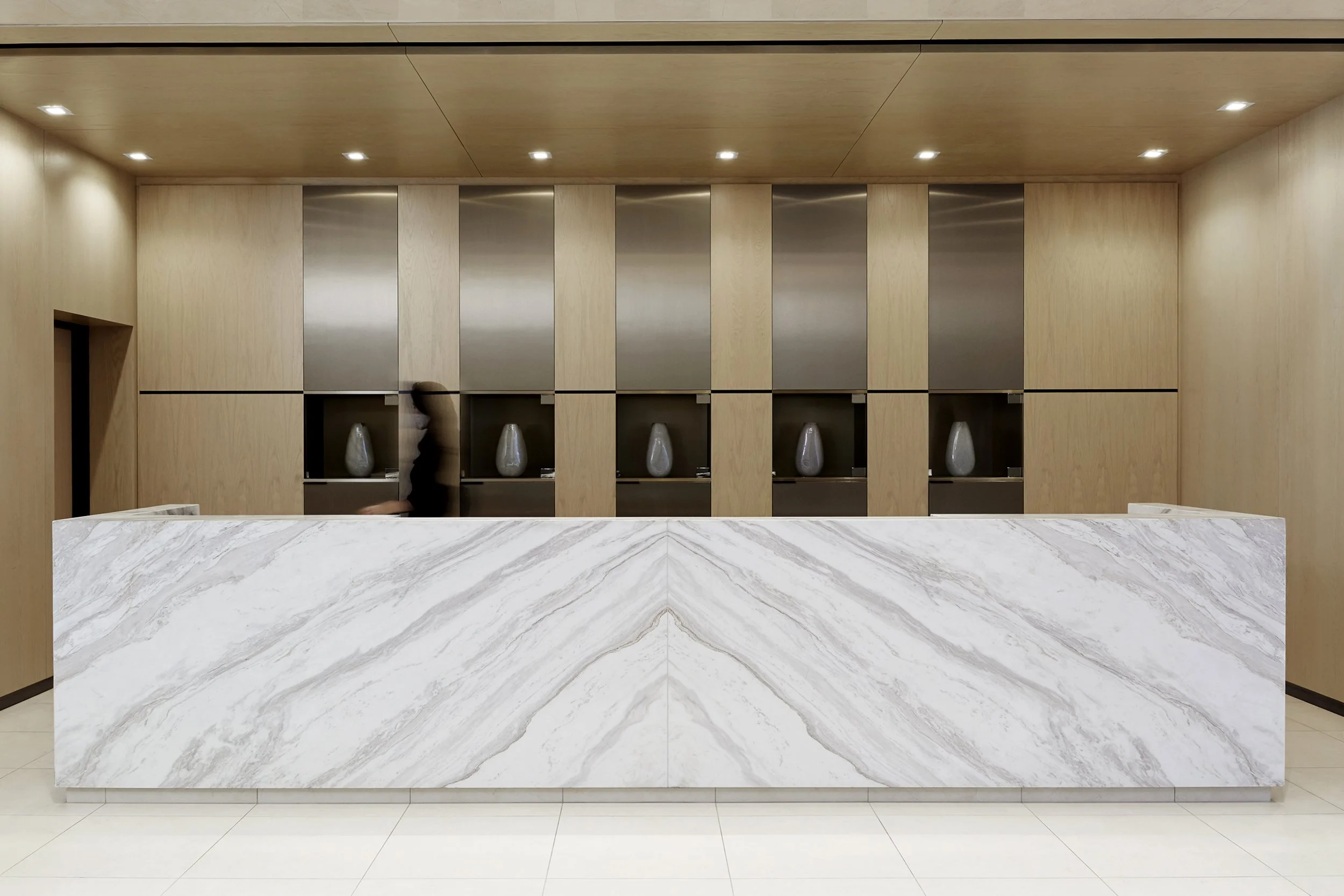 Modern reception desk at Mattamy Homes - J. Davis House, featuring a marble front, set against a wooden wall with metallic panels and decorative vases displayed within recessed wall niches.