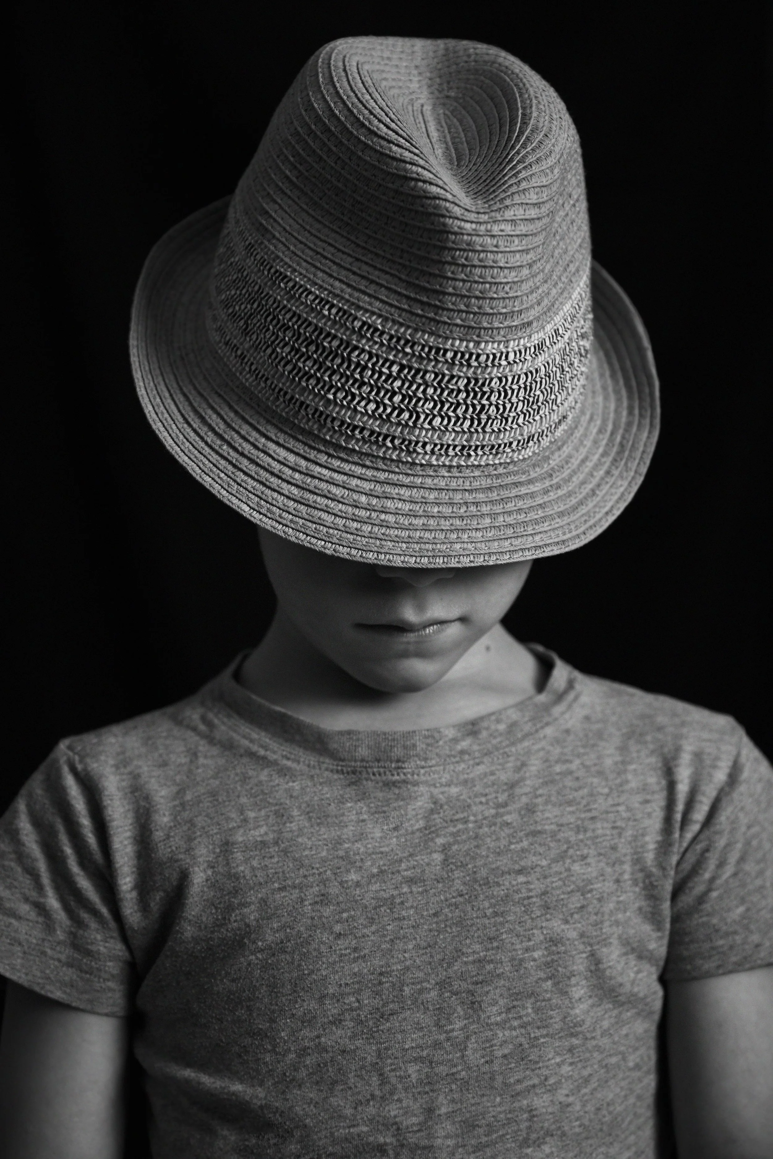 A person wearing a large, textured hat and a plain t-shirt, with their head tilted downward, in black and white against a black background.