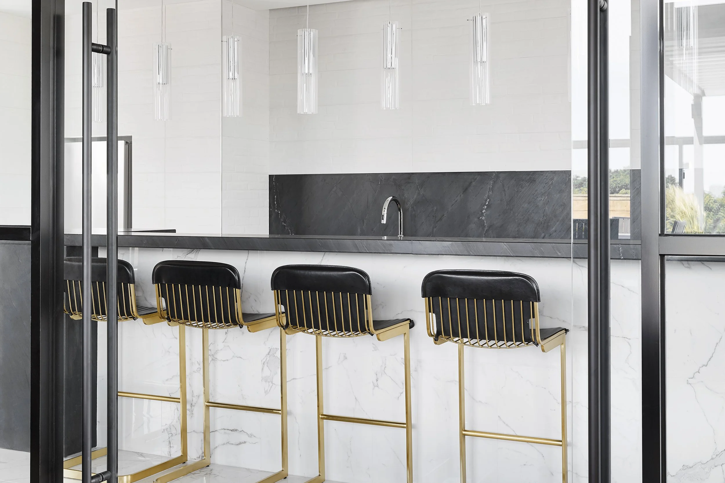 Party room kitchen and bar at Mattamy Homes - J. Davis House Condominium, featuring four black-and-gold chairs, a marble countertop, a white brick wall, and pendant lights in the background.