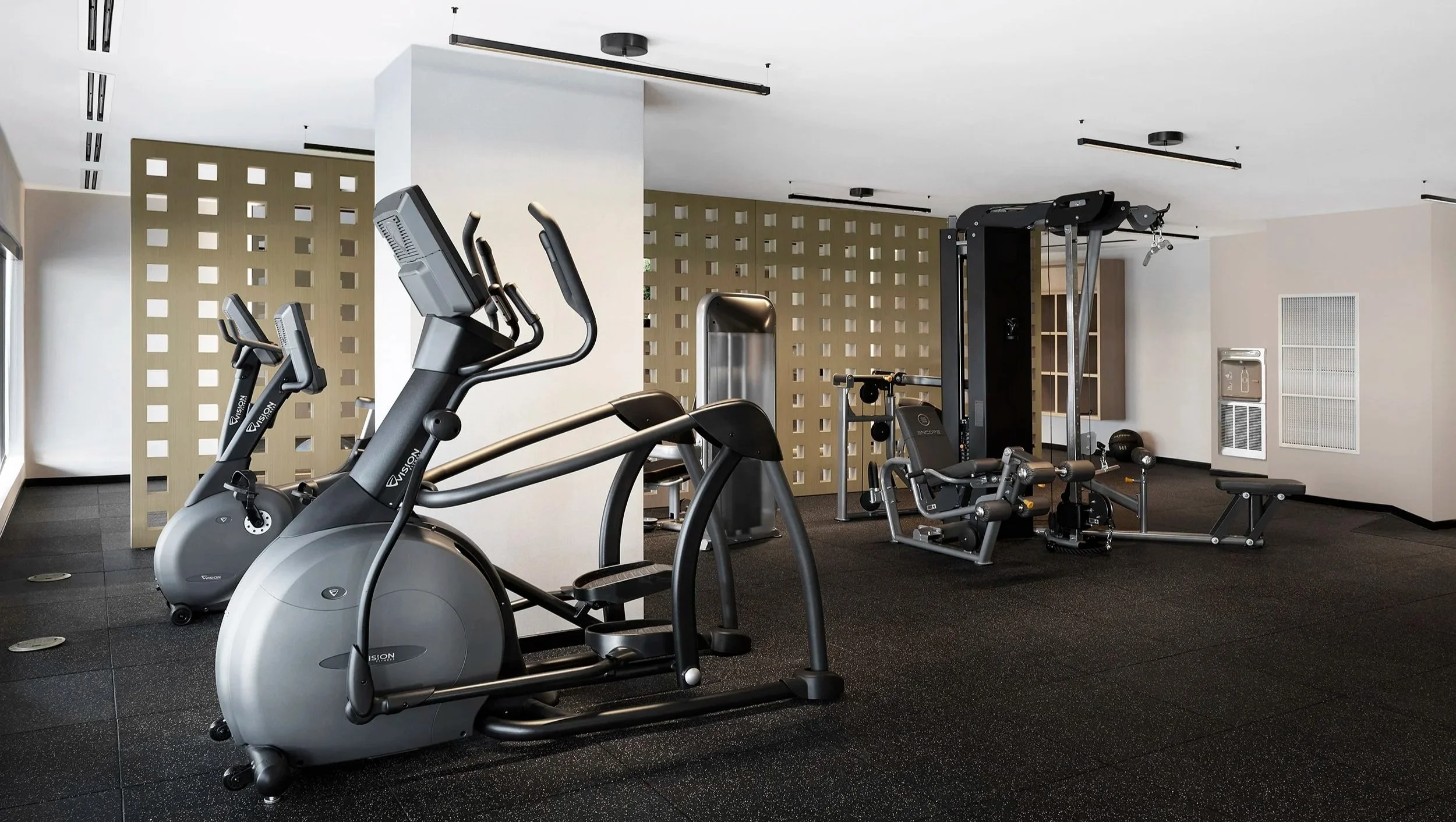 Gym at Station No. 3 Condo in Whitby, with exercise equipment including stationary bikes, elliptical, weight machines, and benches on a black rubber floor, with a patterned room divider and wall-mounted lights.