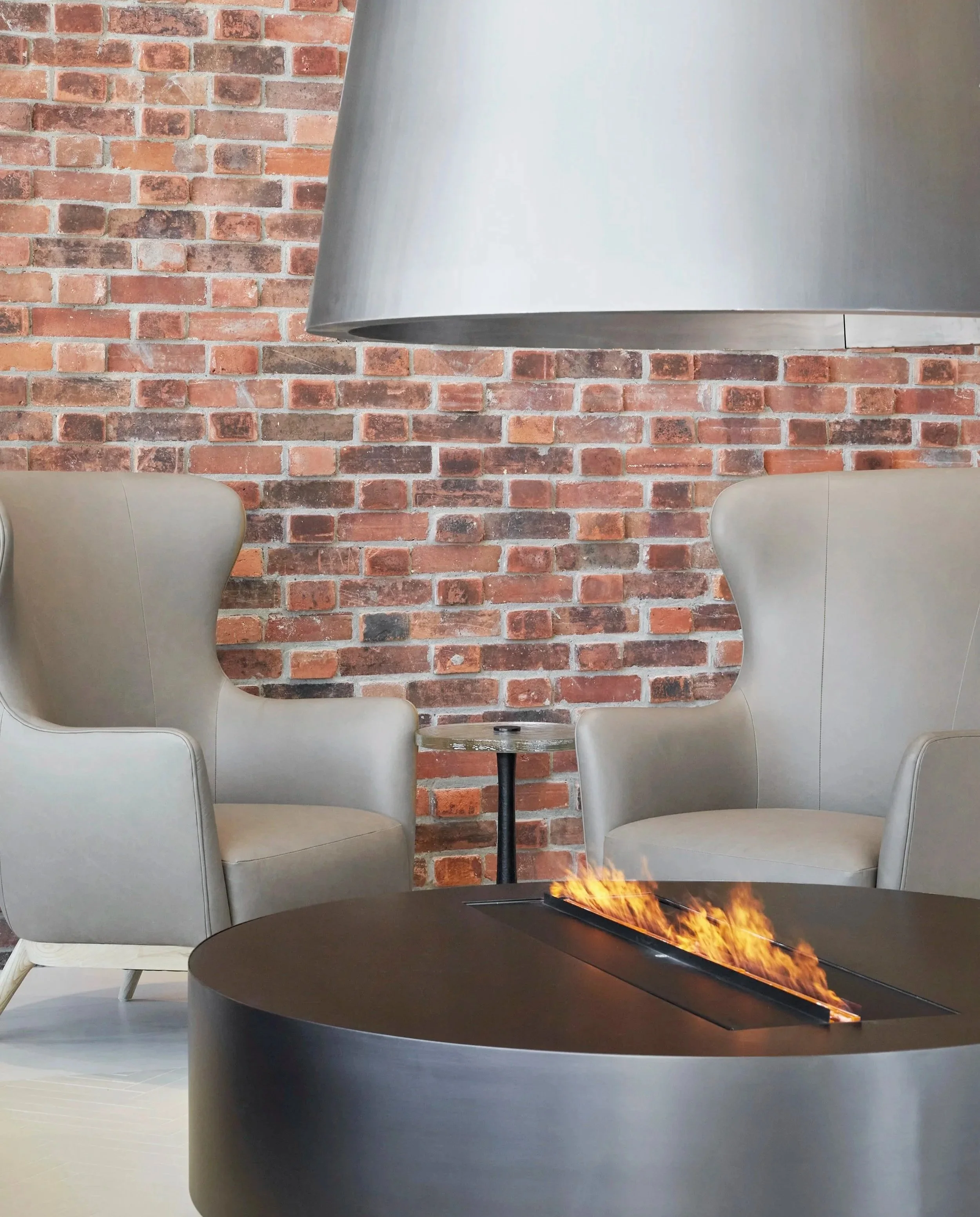 Lobby seating at Station No. 3 Condominiums in Whitby by Brookfield Properties, a metal fire pit table with flames, a small round side table, and a brick wall background.