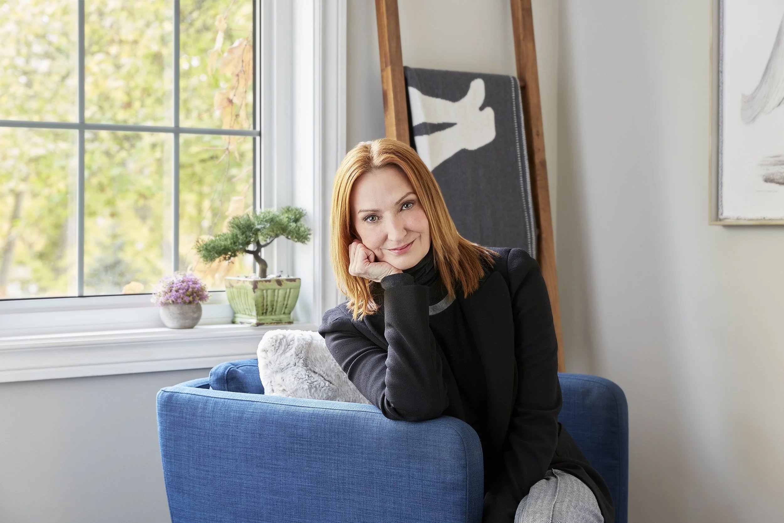 A woman with red hair sitting on a blue chair near a window with green trees outside, resting her chin on her hand and smiling at the camera.