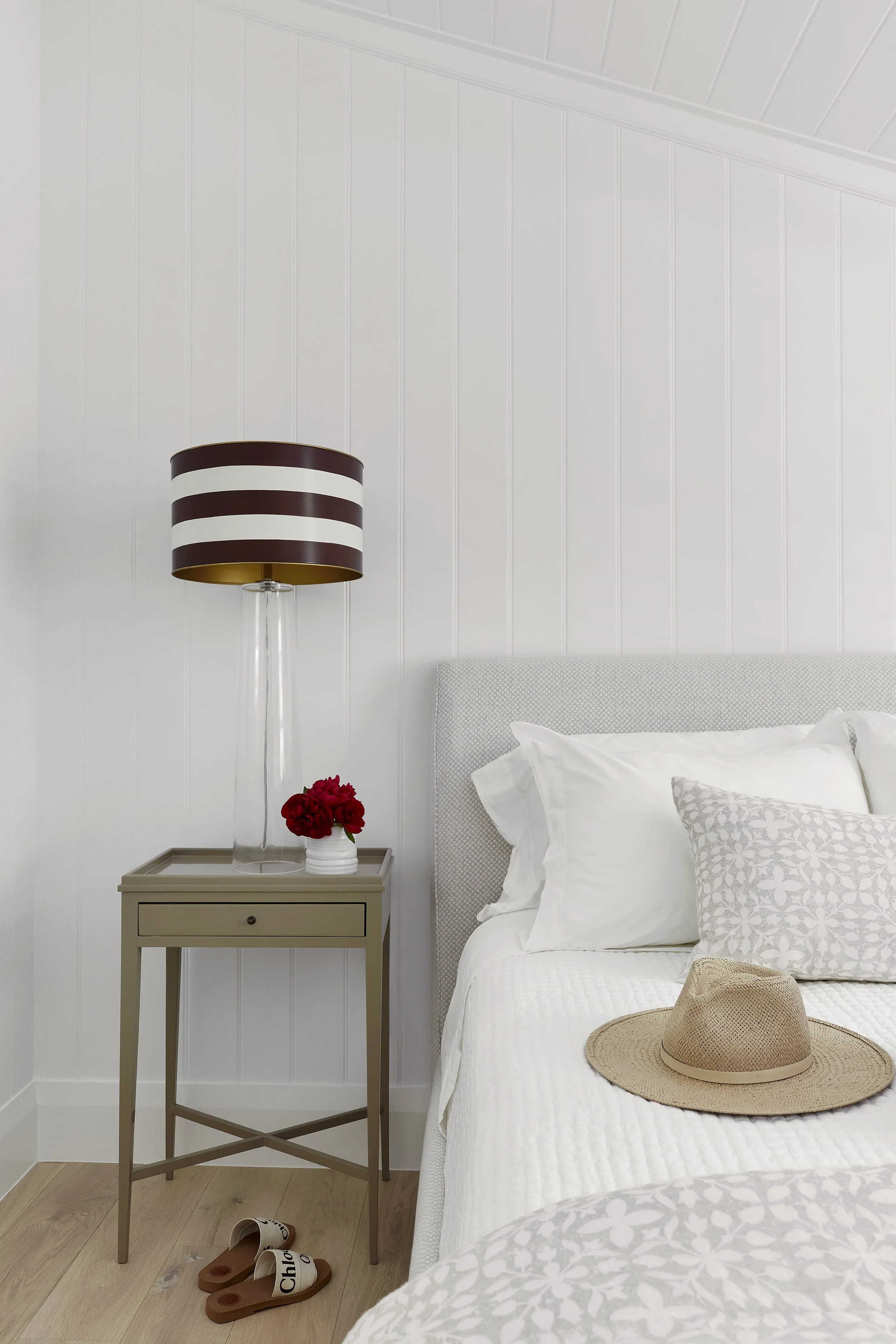 A bedroom with a bedside table, a lamp with a striped shade, a small vase of red flowers, a bed with white bedding and decorative pillows, and a straw hat on the bed. There are slippers on the wooden floor.
