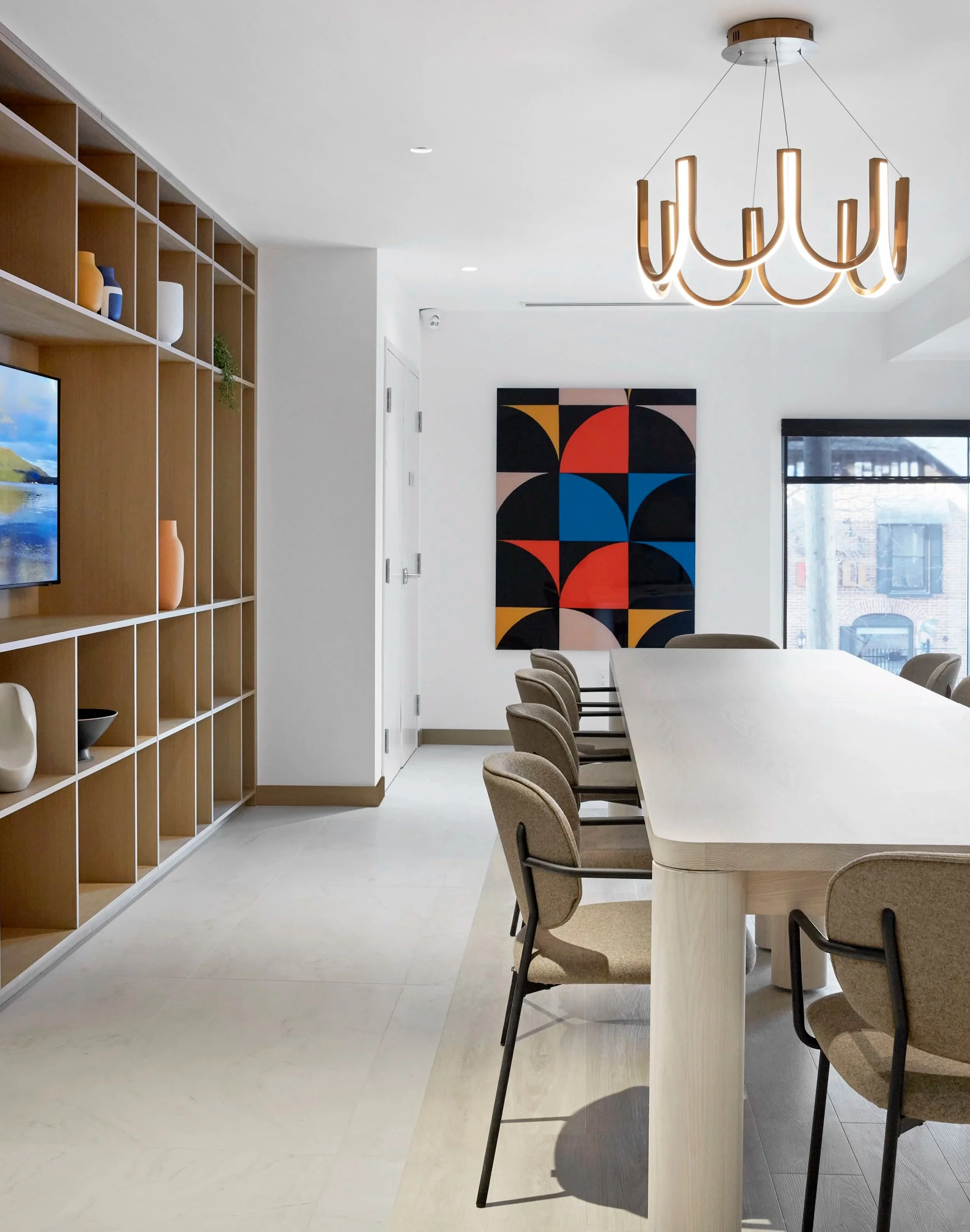 Conference room co-working space at Station No. 3 Condominiums in Whitby by Brookfield Properties, featuring a conference table, chairs, a geometric colourful wall artwork, windows with blinds and a contemporary chandelier.