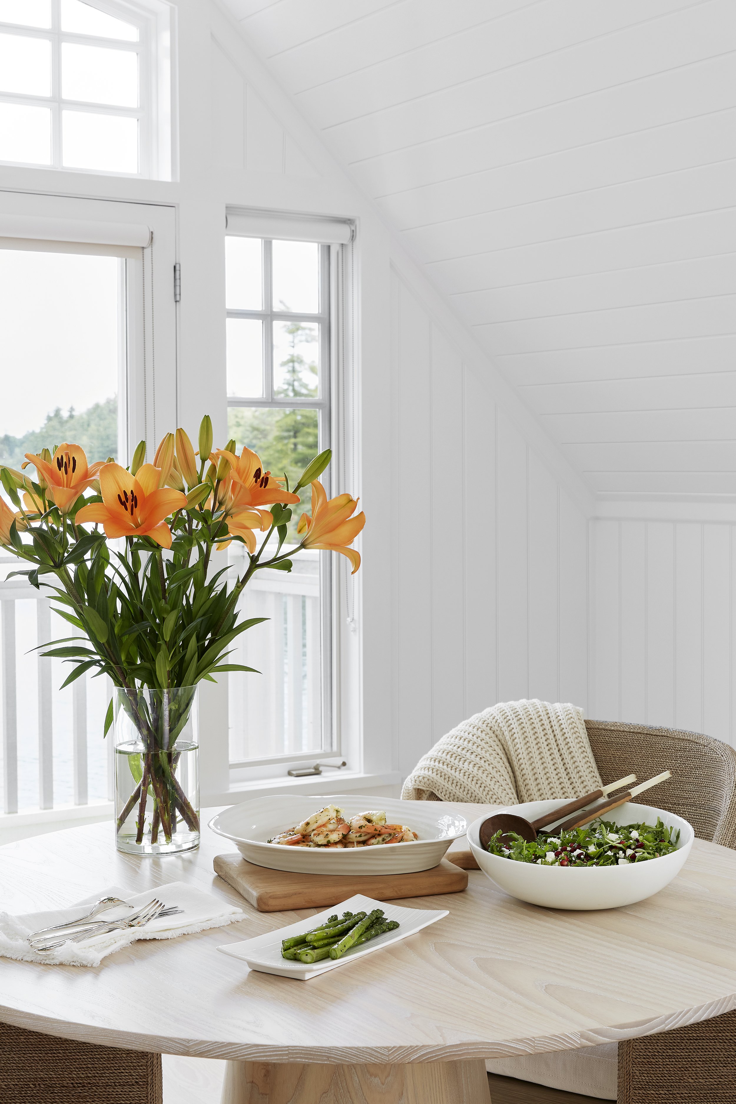 A dining table set near windows with a vase of orange lilies, a bowl of salad, a plate of pasta, a small dish of asparagus, and utensils.