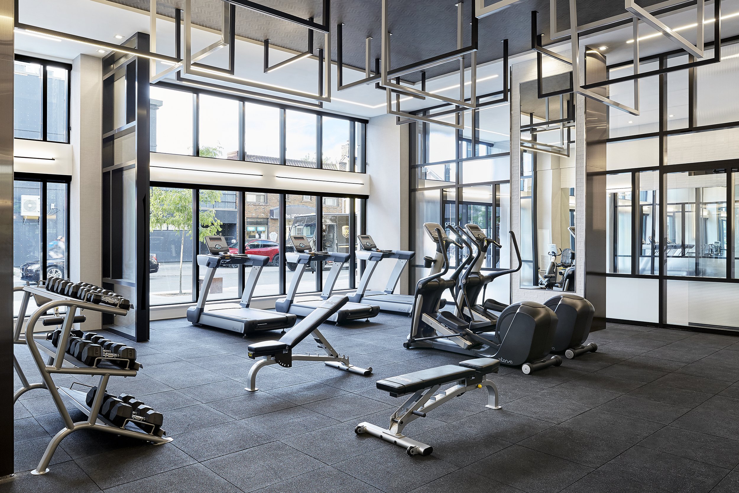Spacious gym at Mattamy Homes - J. Davis Condominium, featuring large floor-to-ceiling windows, treadmills, ellipticals, free weights, workout benches, and decorative ceiling elements in a bright, open space.