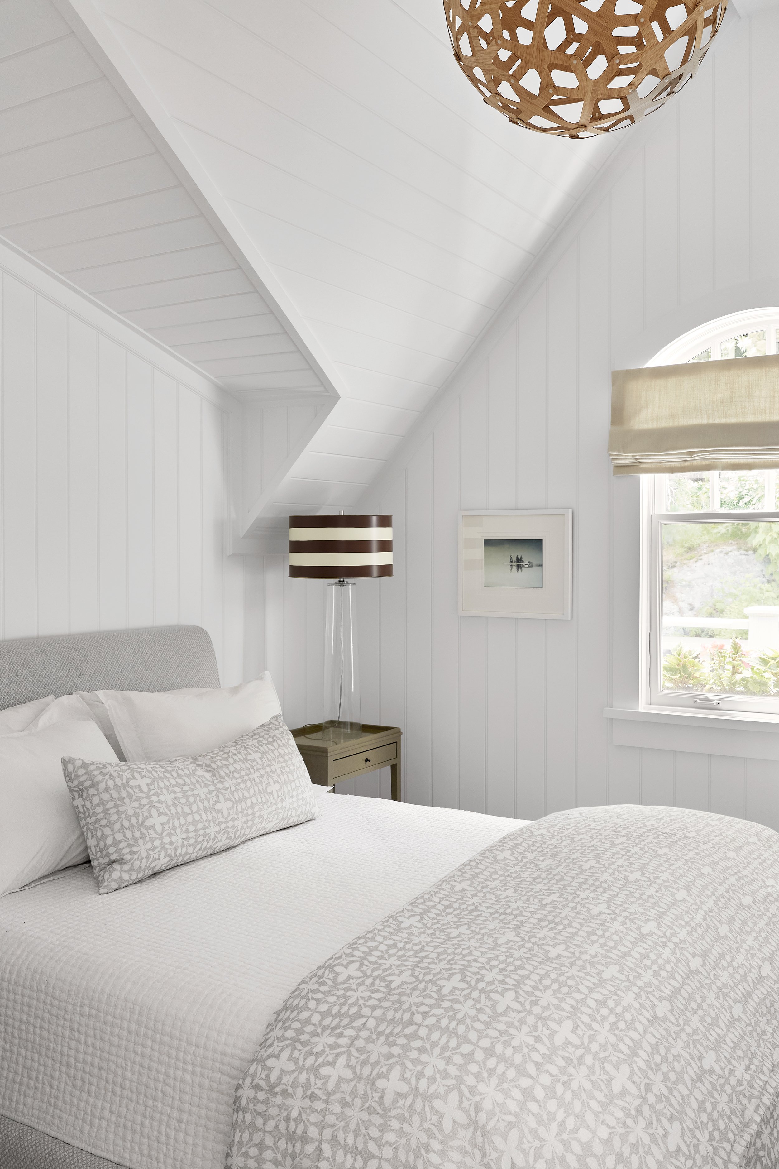 A cozy bedroom with a sloped white ceiling and white paneled walls, a bed with white linens and a decorative gray patterned pillow, a small nightstand with a glass lamp with brown and white stripes, a window with a beige Roman shade, a framed picture