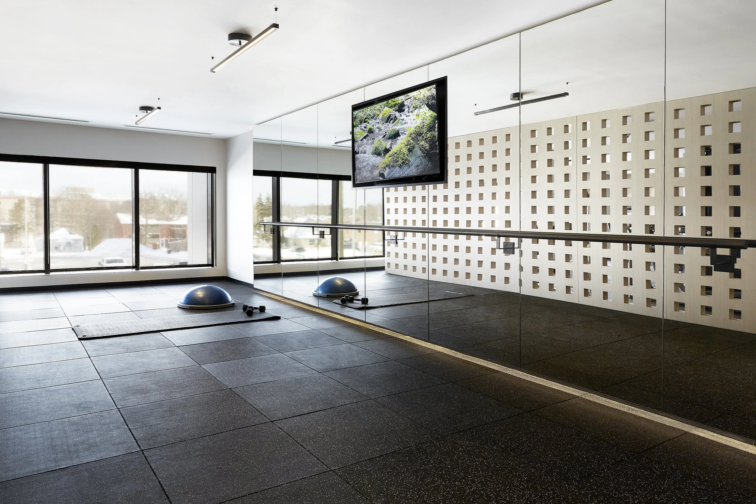 Yoga and fitness room at Station No. 3 Condominiums in Whitby, featuring black rubber flooring, two blue half-dome balance trainers, small black dumbbells, large windows, a wall mirror, a mounted TV, and a ballet barre along the mirrored wall.
