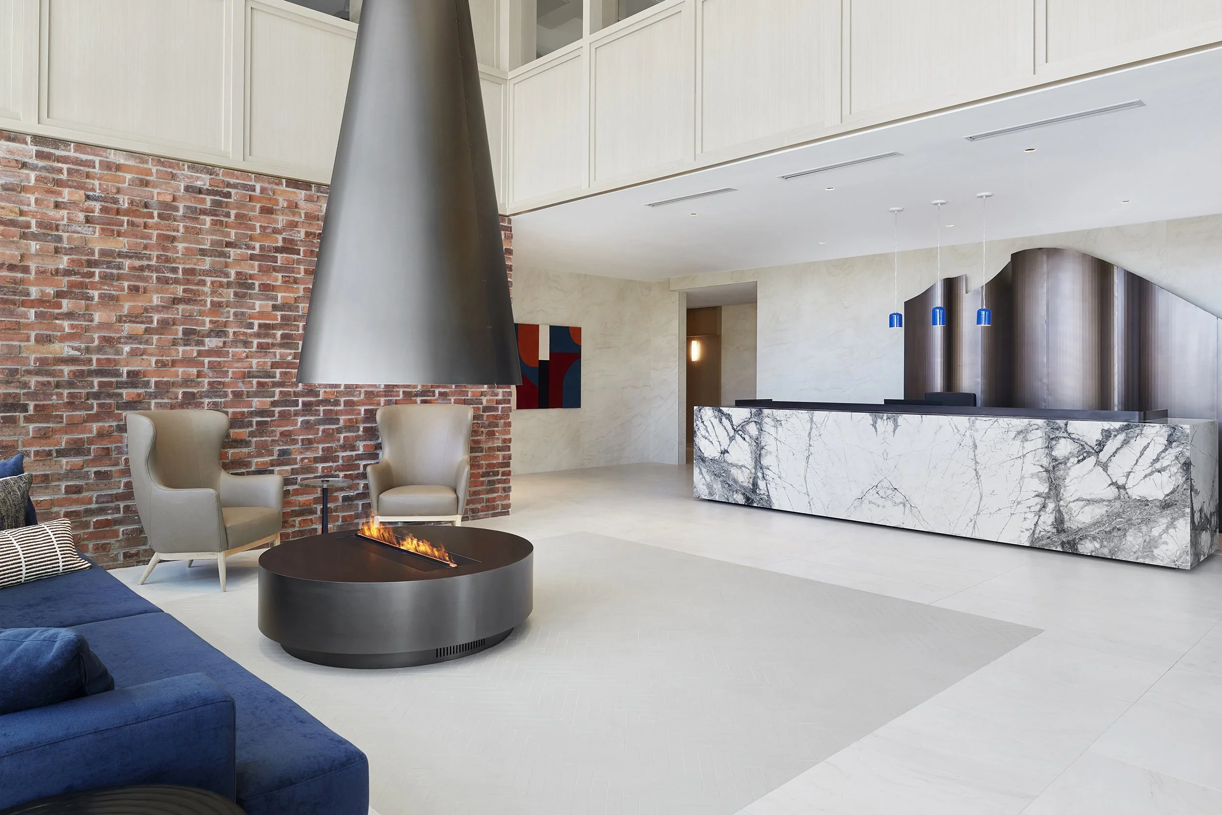 Modern lobby at Station No. 3 Condominiums in Whitby by Brookfield Properties, a restored historic fire station featuring exposed brick walls, beige armchairs, a circular fireplace, a blue sofa, and a marble reception desk with pendant lighting.