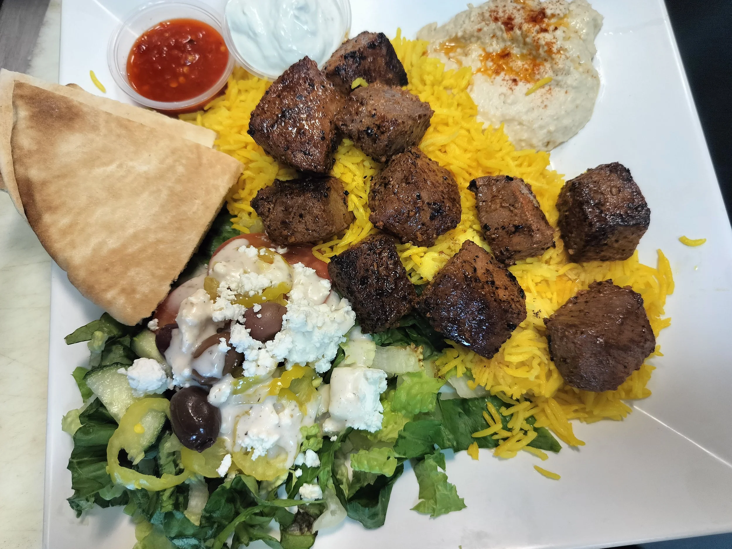 A plate of lamb kabab It includes grilled lamb cube cubes, yellow rice, a piece of flatbread, a small bowl of red sauce, a dollop of creamy white sauce, and a serving of salad with feta cheese, olives, cucumbers, and dressing.