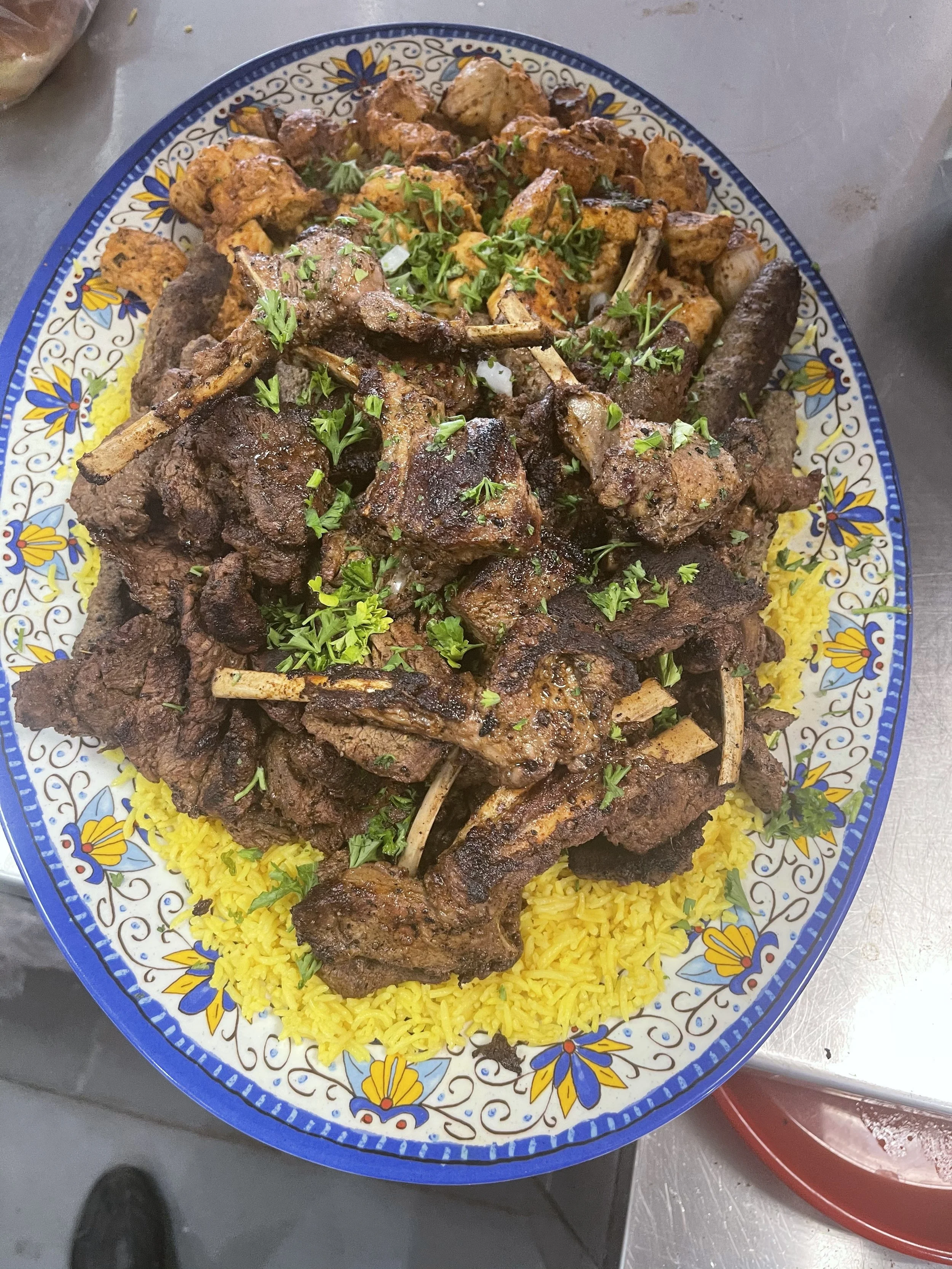 A colorful plate of grilled beef and lamb kabab and chops served on a bed of yellow rice, garnished with chopped fresh herbs, on a decorative ceramic plate.