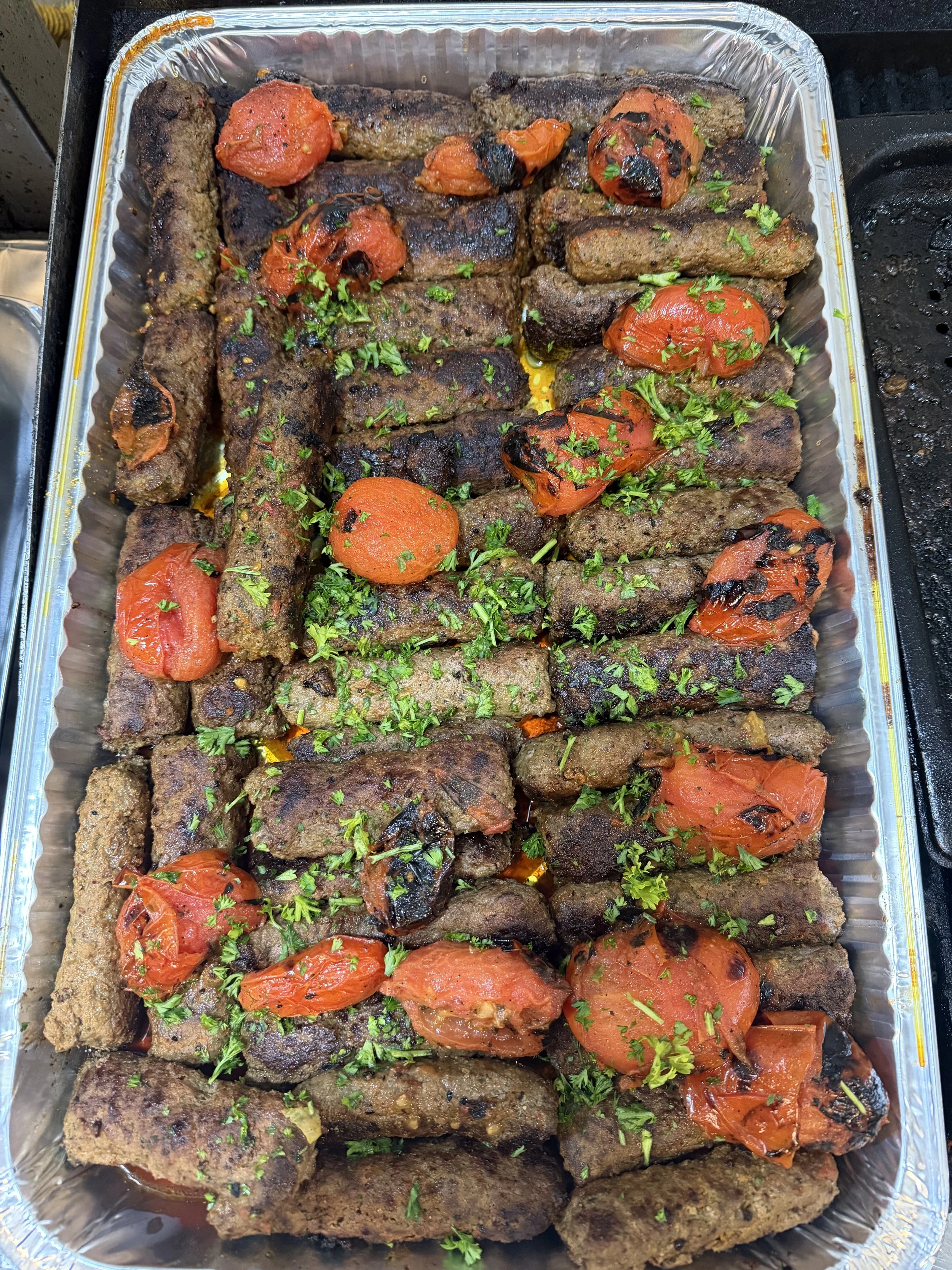 Tray of cooked kebabs garnished with chopped herbs and grilled tomatoes.