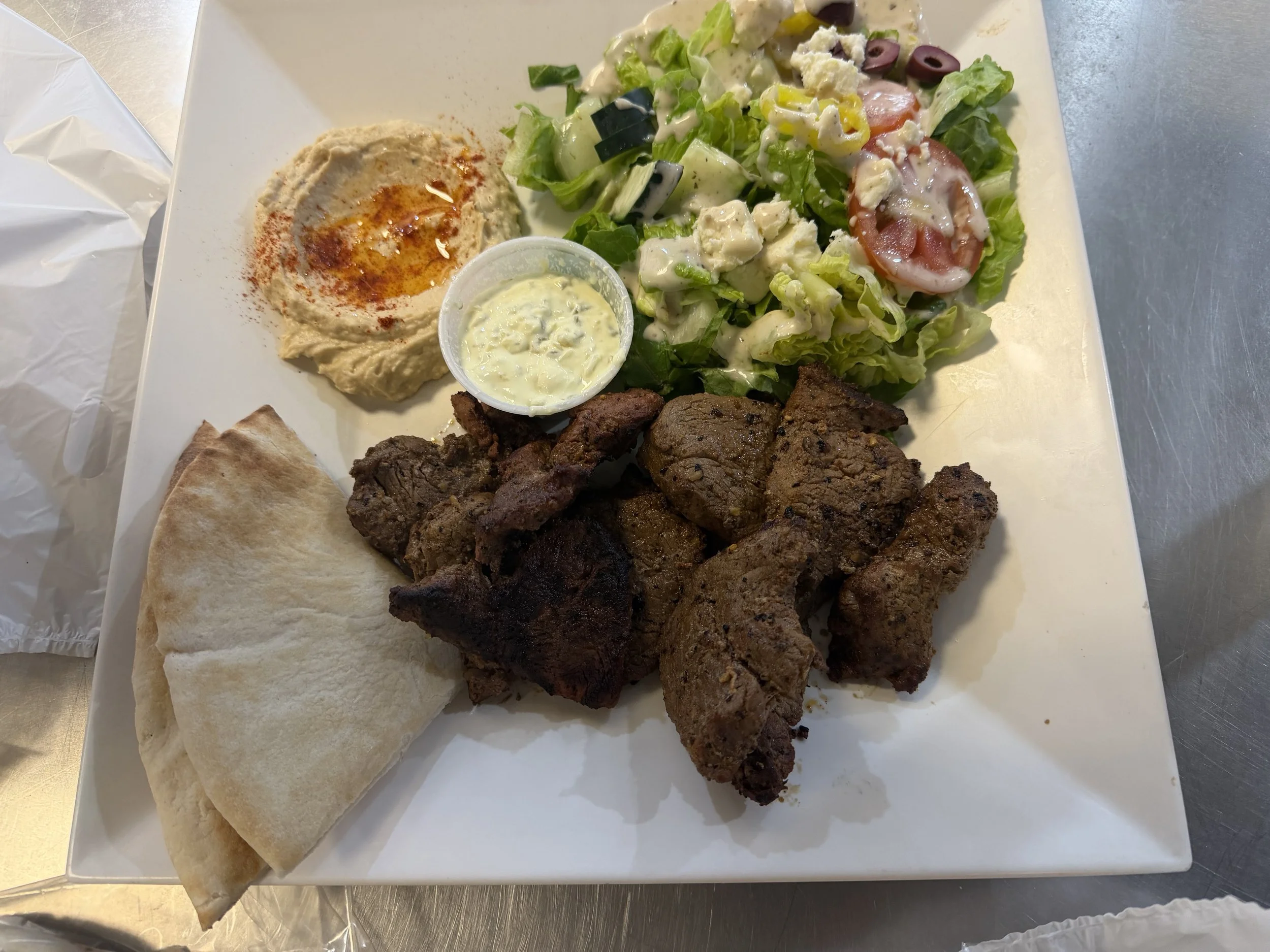 A plate with grilled meat, a side of hummus garnished with paprika and olive oil, a small bowl of creamy salad, a piece of pita bread, and a mixed salad with lettuce, tomatoes, cucumbers, olives, and feta cheese.