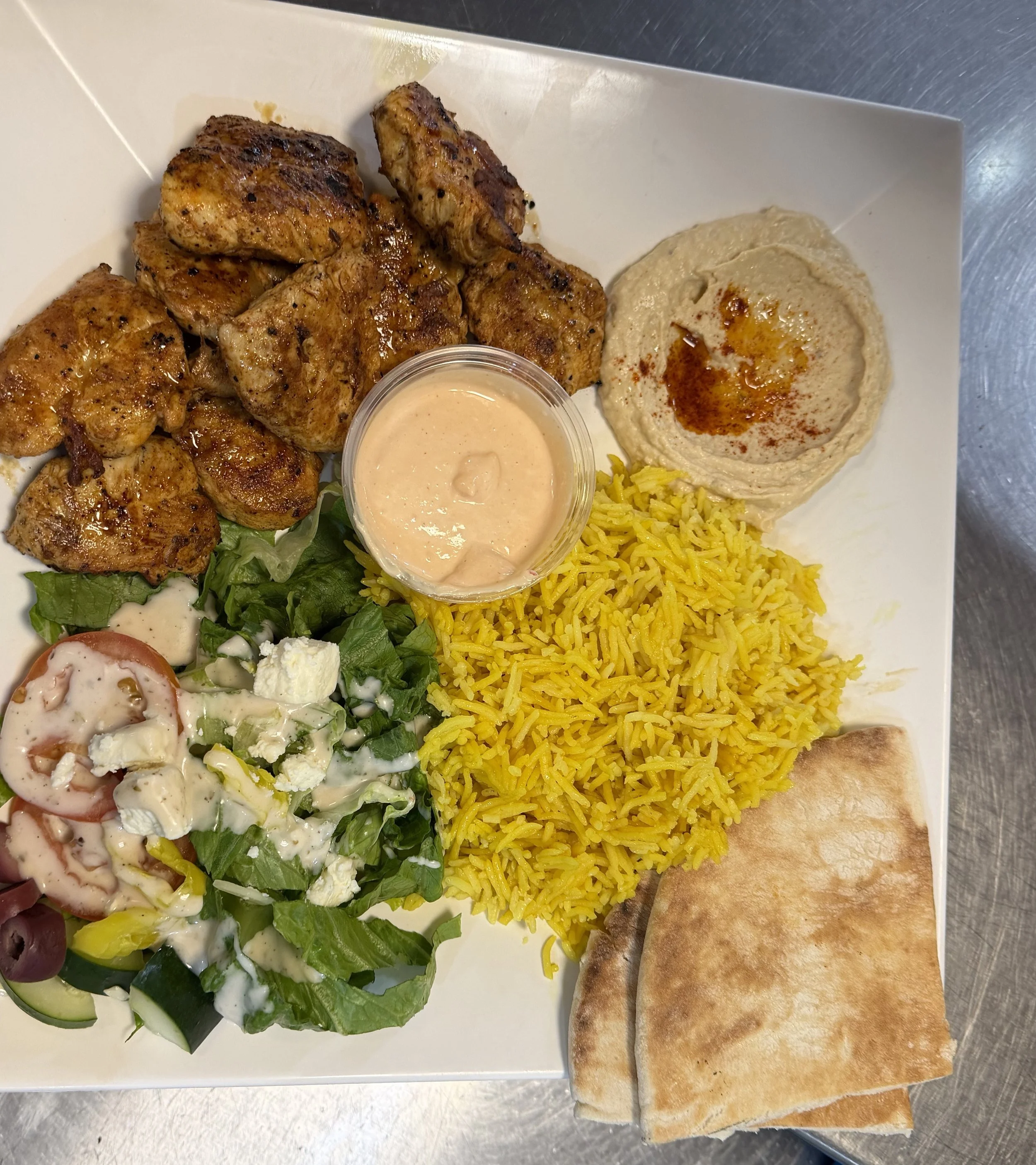 Plate of grilled chicken kabab, yellow rice, creamy hummus with paprika, fresh salad with tomato, cucumber, feta cheese, and dressing, with pita bread on the side.