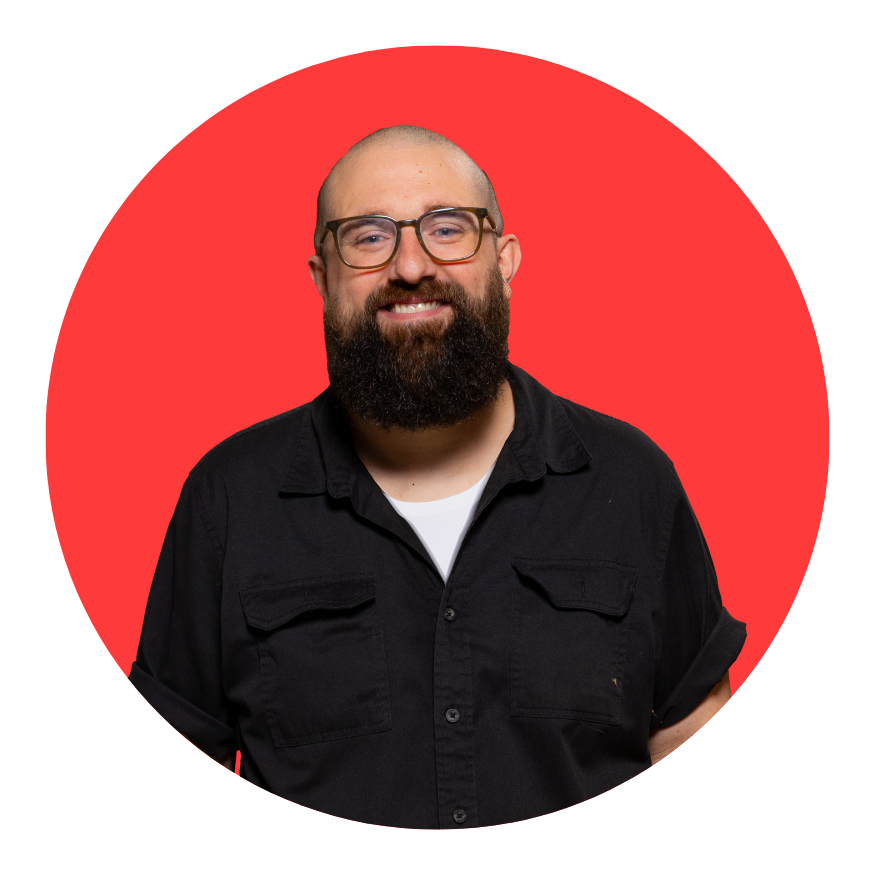 A smiling man with a beard, glasses, and a shaved head, wearing a black button-up shirt over a white T-shirt, standing in front of a red circular background.