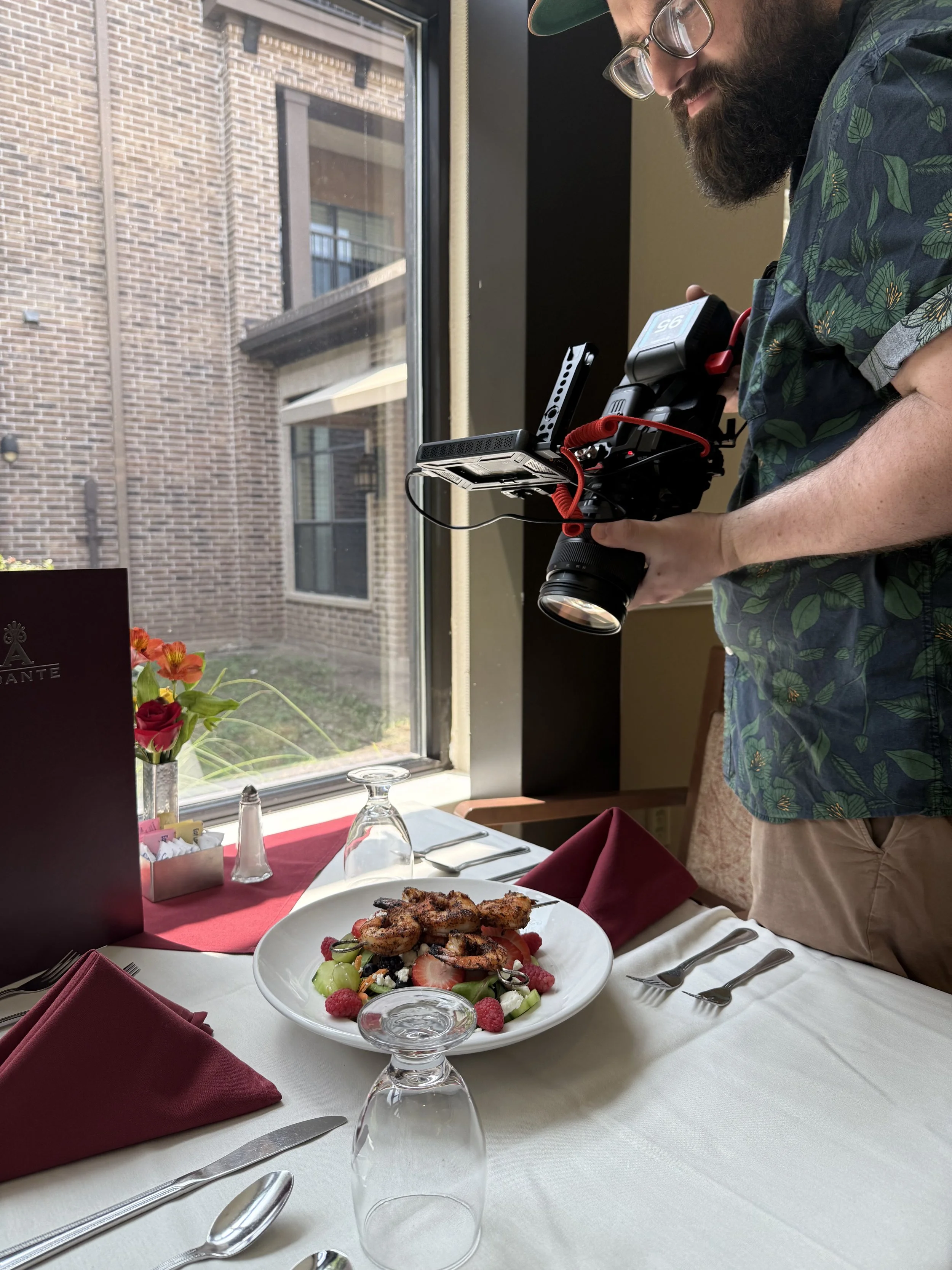 A man with a beard, wearing glasses and a green hat, is filming a plate of salad and grilled chicken on a white tablecloth at a restaurant with large windows, brick buildings, and a small garden outside.