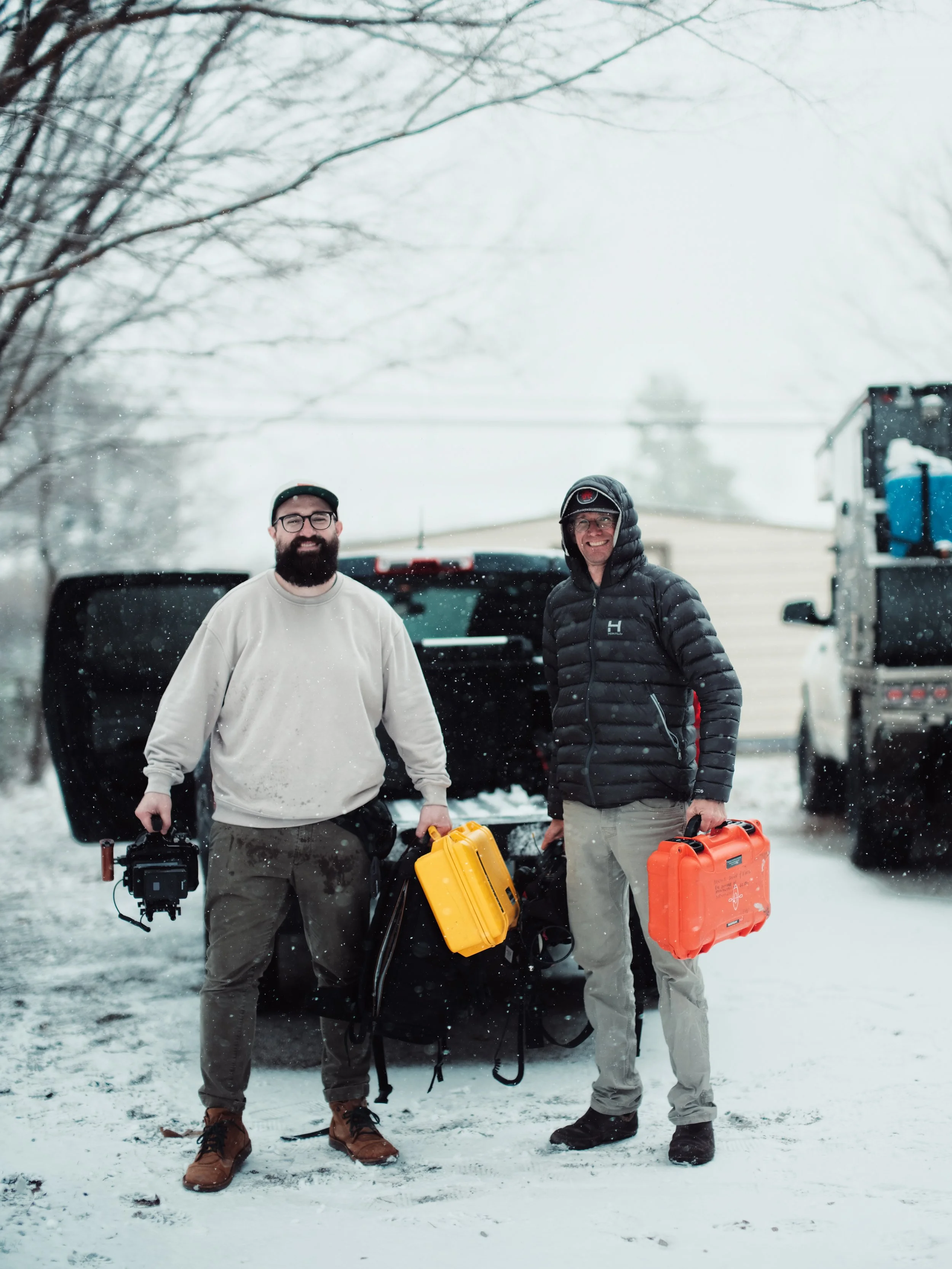Two men standing in a snowy outdoor area, each holding a yellow or orange emergency medical kit, with vehicles and snow-covered trees in the background.