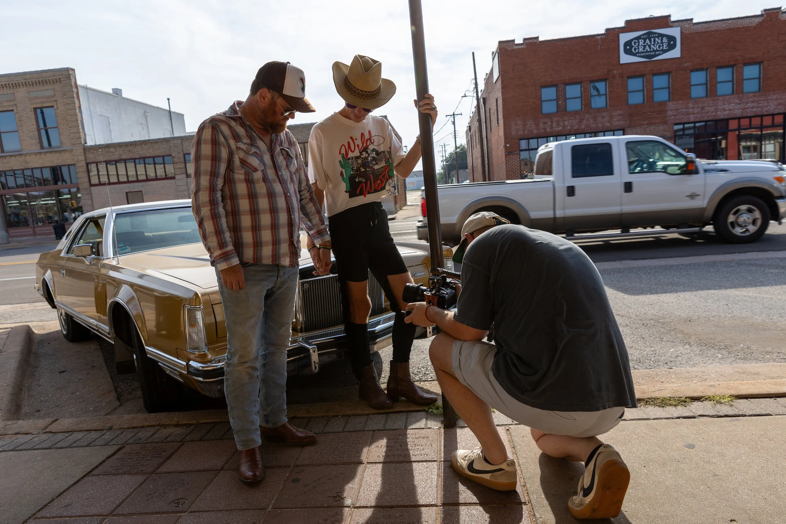 Group of four people working together outdoors at a street corner during the daytime. One person is wearing a plaid shirt and a trucker hat, standing next to a vintage gold-colored car. Another individual, wearing a wide-brimmed hat and a graphic T-shirt, is holding a pole. A third person, wearing a cap, is crouched down examining something on the ground. There is a building with a sign that reads 'GRAIN & GRANGE' in the background, and a white pickup truck is passing by on the street.