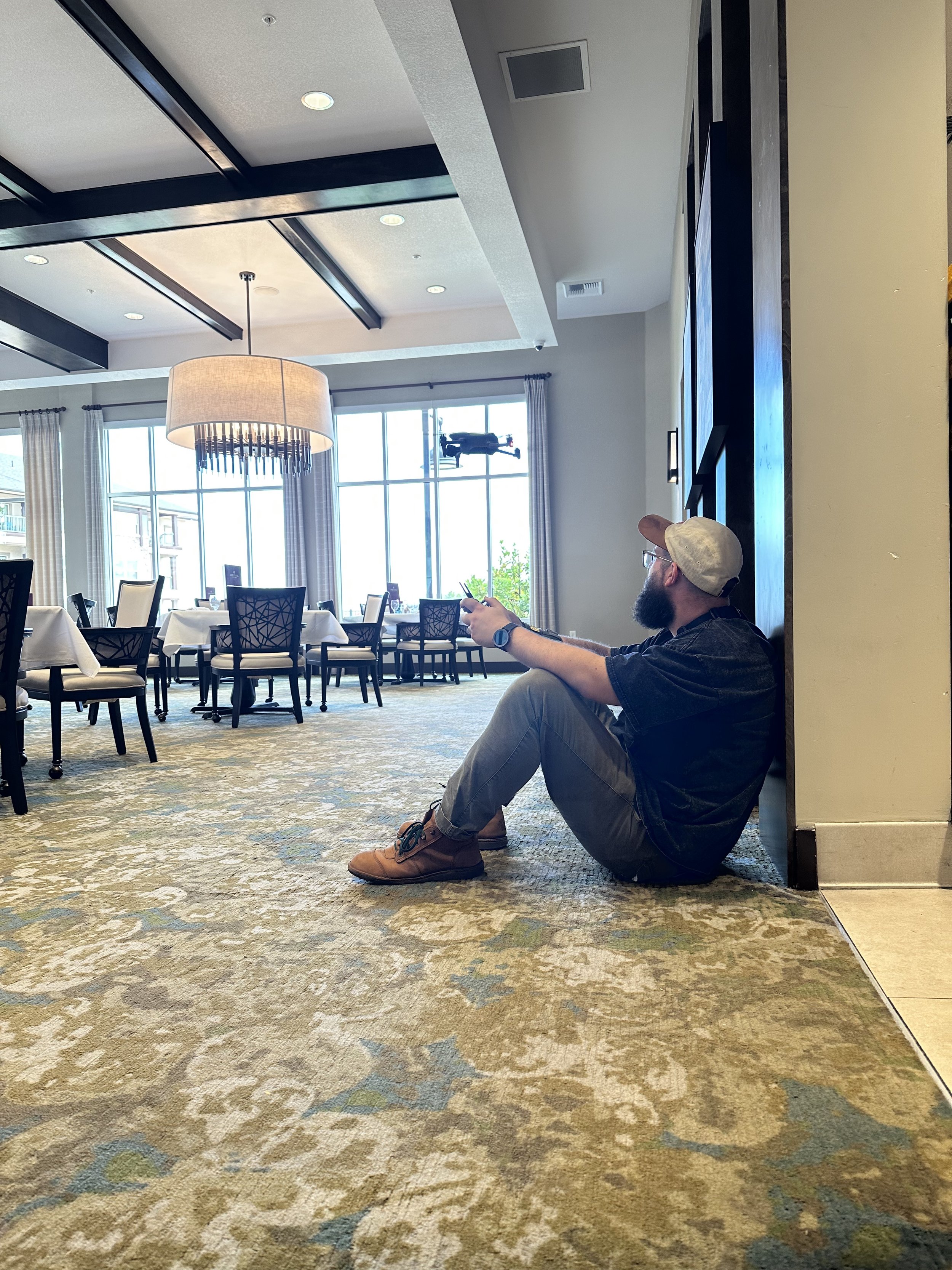 A man with a beard, glasses, and a beige cap is sitting on the floor in a dining area, holding a phone, with a drone flying outside through large windows behind him. The room has white curtains, a chandelier, and round dining tables with chairs.