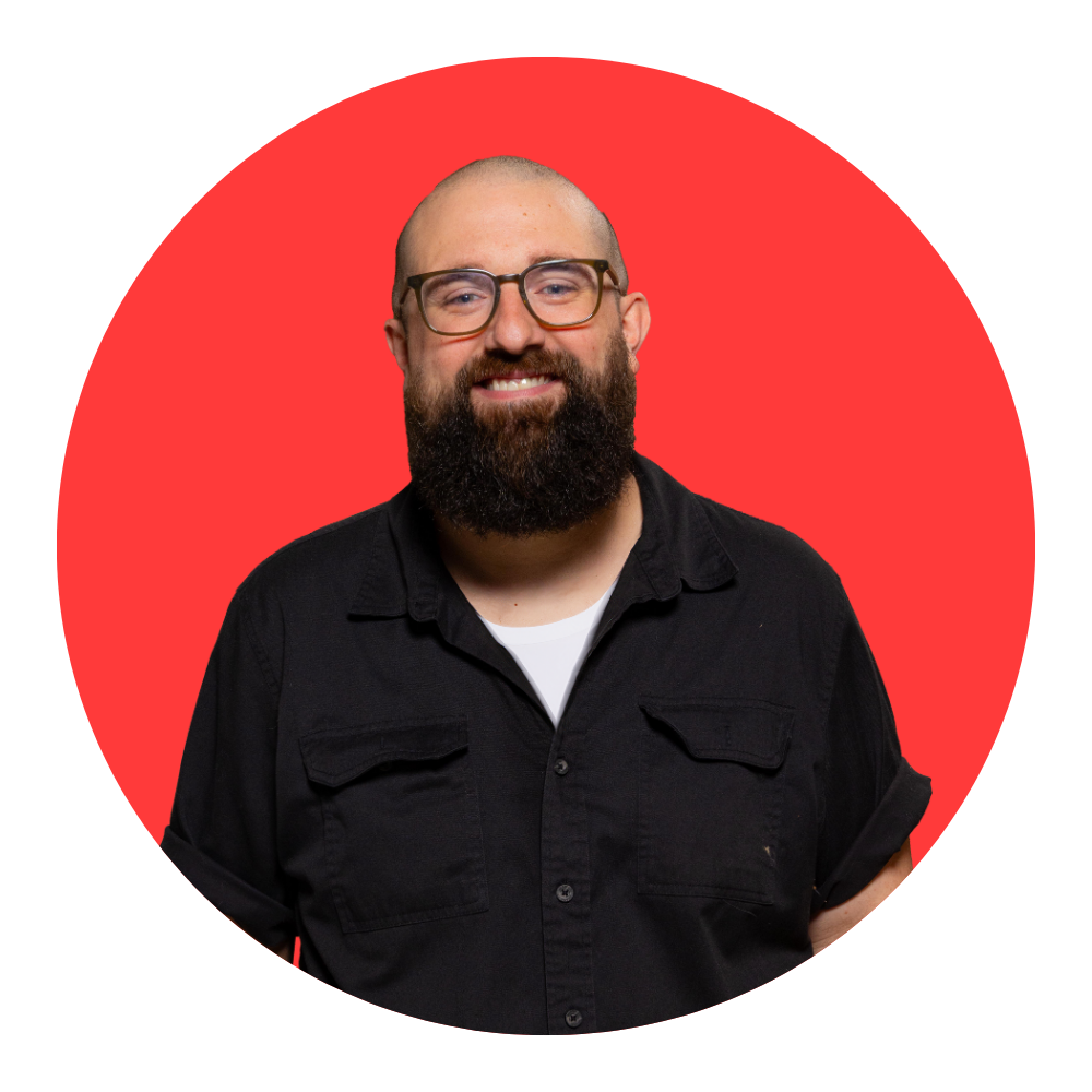 A man with a beard, glasses, and a smile, wearing a black shirt, standing against a red circular background.
