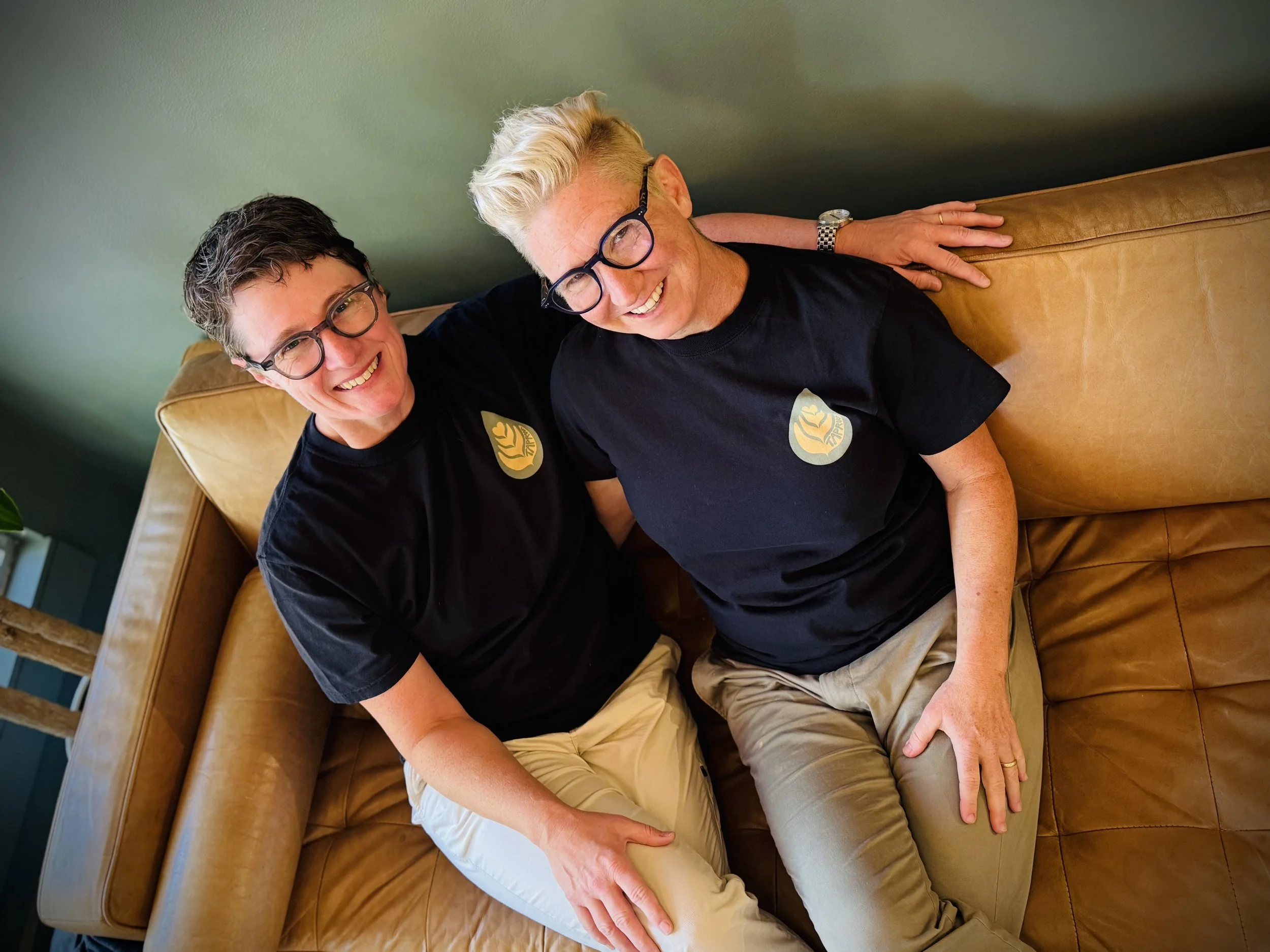 Owners, Ashley and Alison, in matching drip t-shirts sitting and smiling at the camera