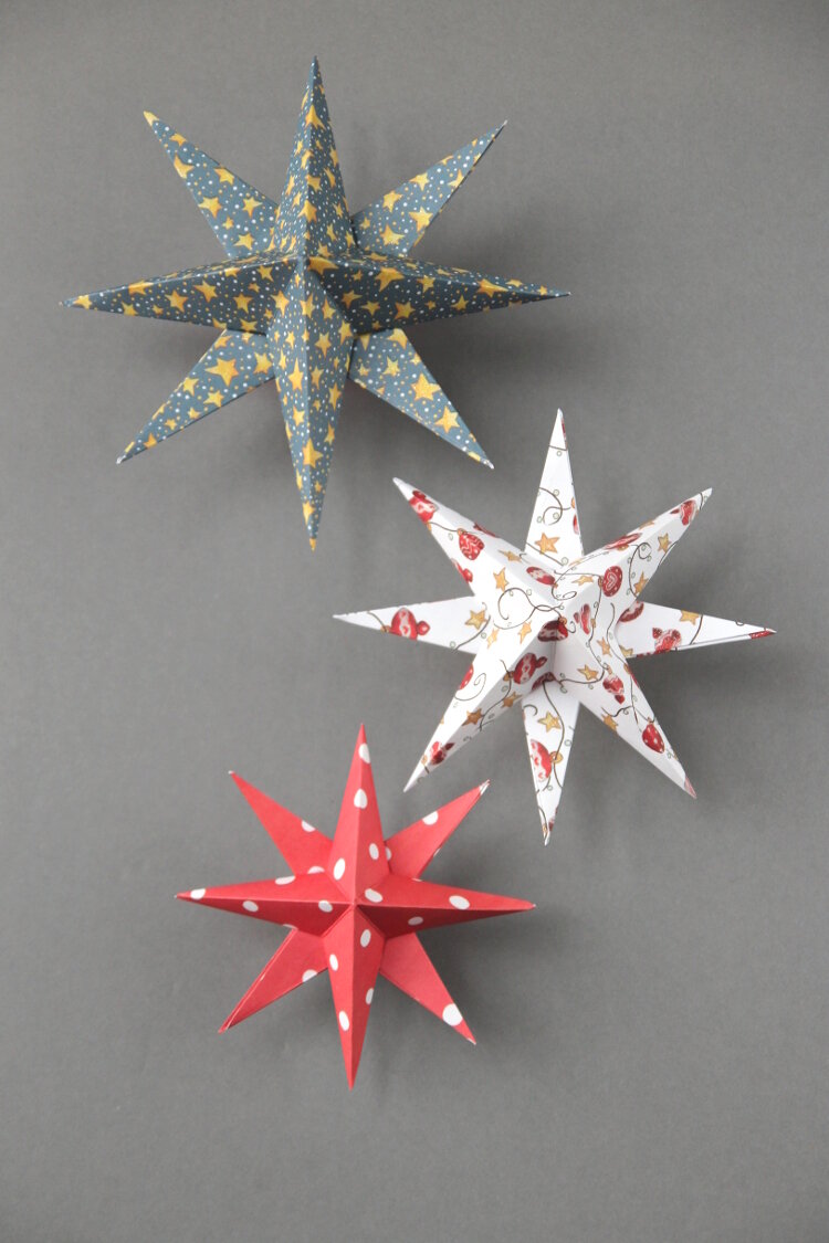 Three 8 pronged decorative paper stars dangle in front of a gray background.  One is gray and yellow, another is red, yellow, and white, and the third is red and white with polka dots.