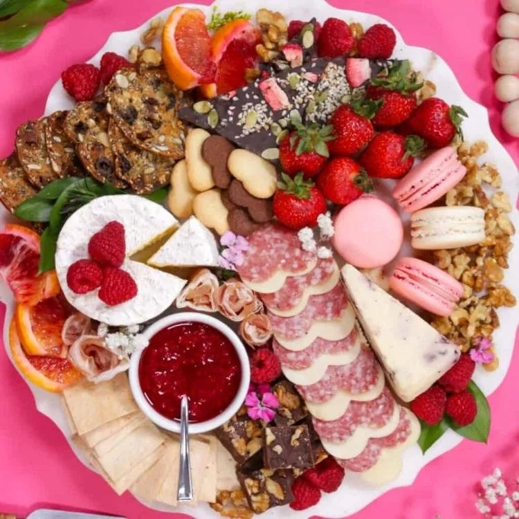 round plate on pink background filled with heart shaped meat, cheese, biscuits, strawberries and macarons